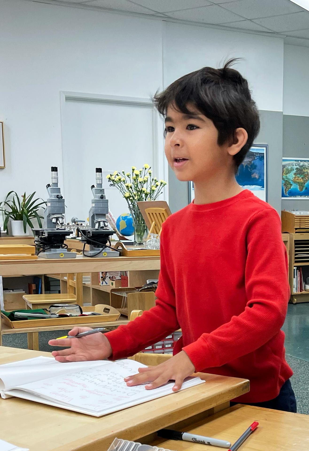 Child in red shirt standing at a desk with an open notebook