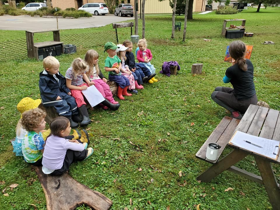 Person sitting on ground facing group of children sitting on grass