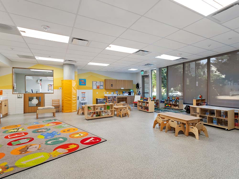 Empty classroom with small wooden tables and chairs, colorful rug, and large windows