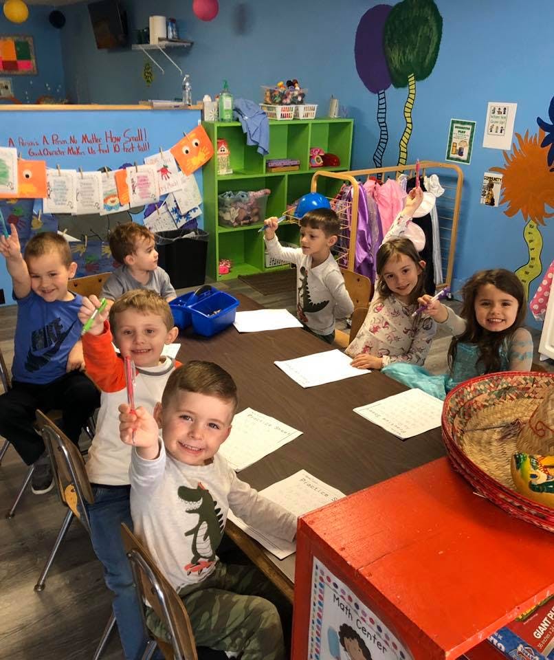 Children sitting at tables in a classroom, some holding up pencils