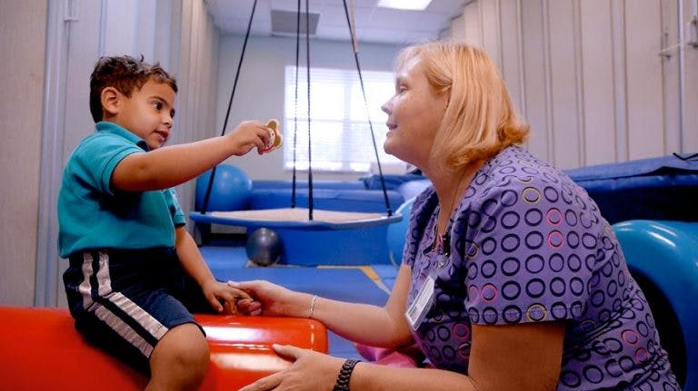 Child sitting on red equipment reaching for person in a therapy room