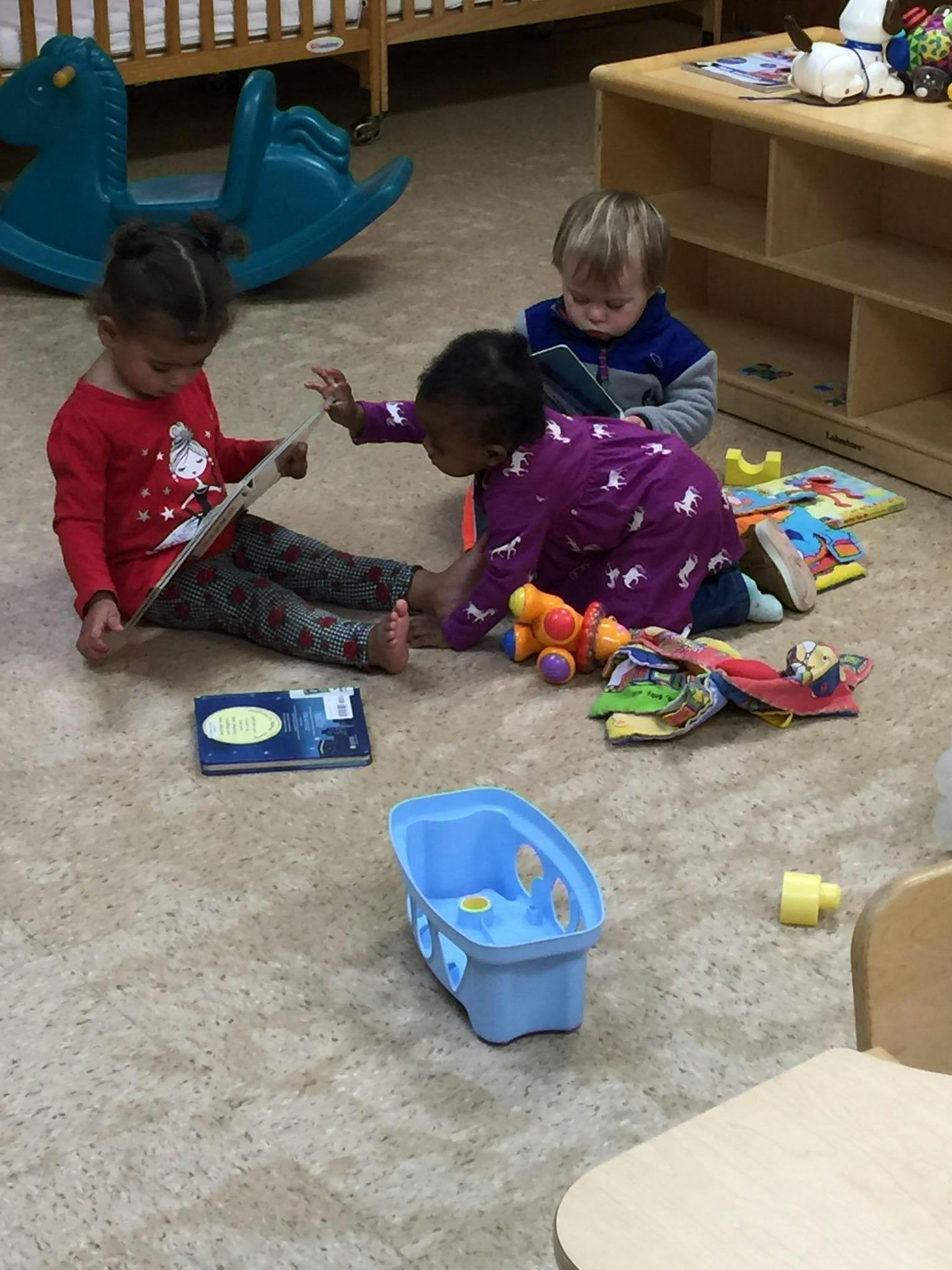 Three children sitting on the floor with books and toys in a classroom
