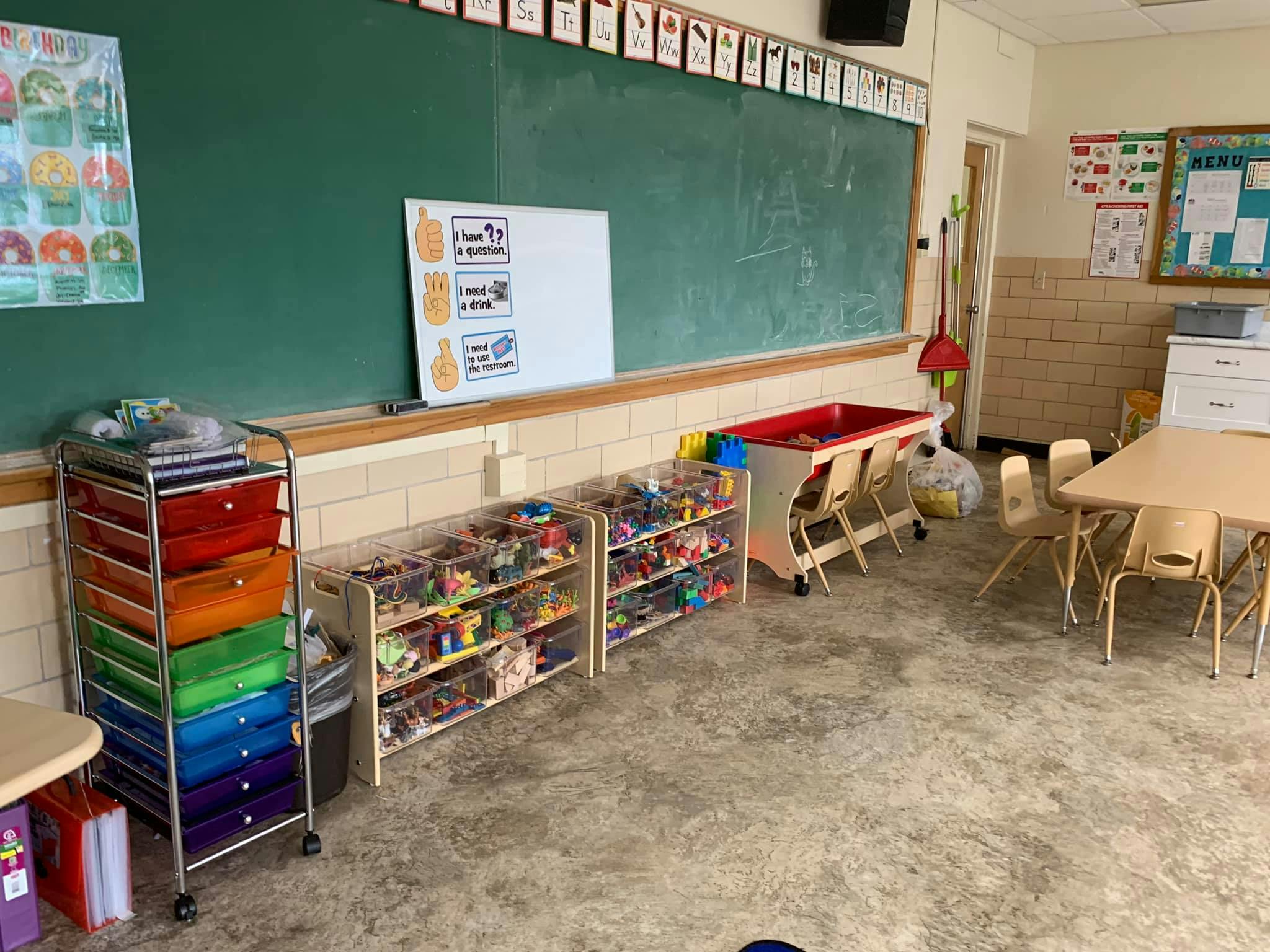 Classroom with a green chalkboard, storage shelves, and small tables