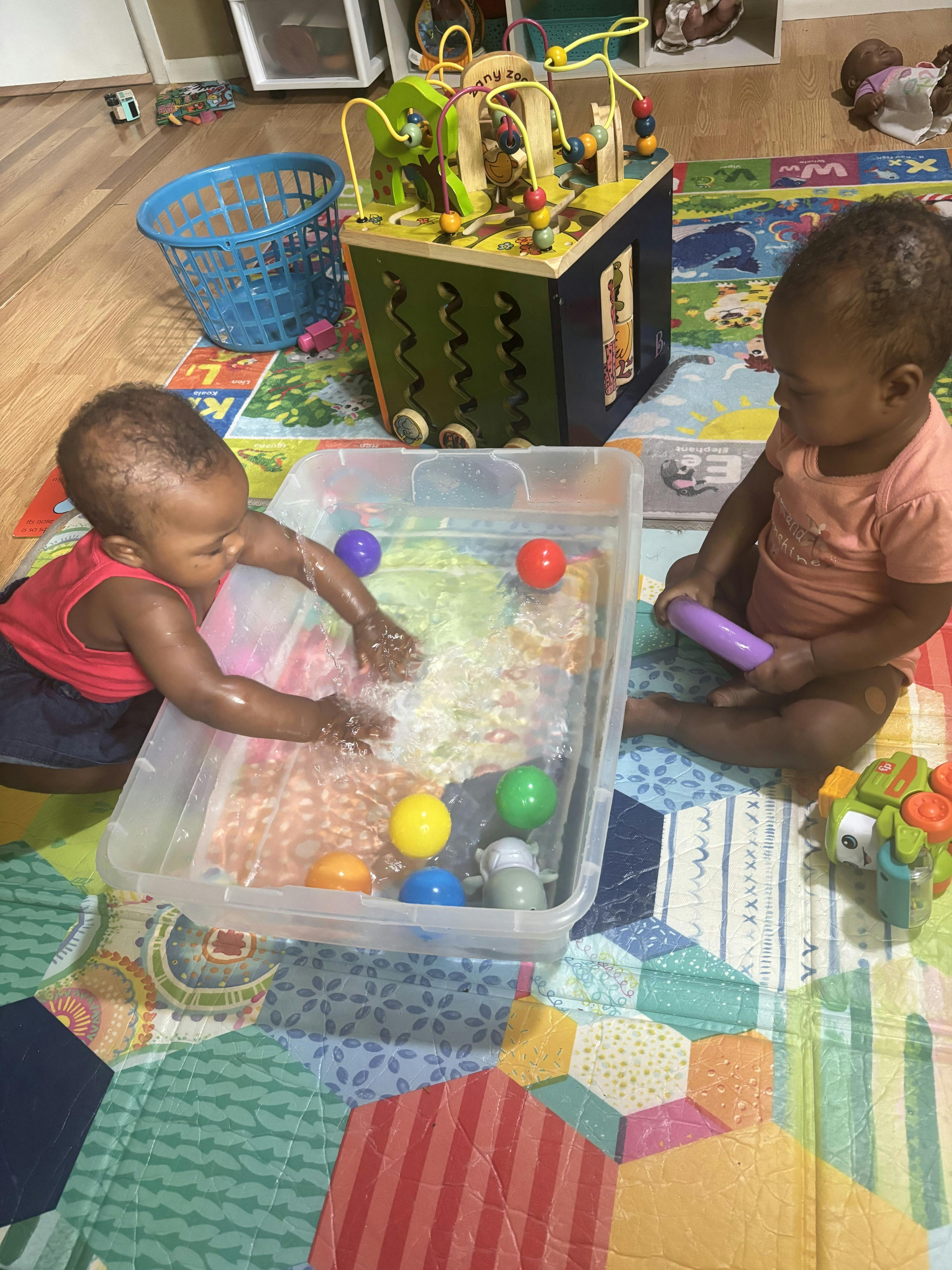 Two children playing with water and plastic balls in a bin on a play mat