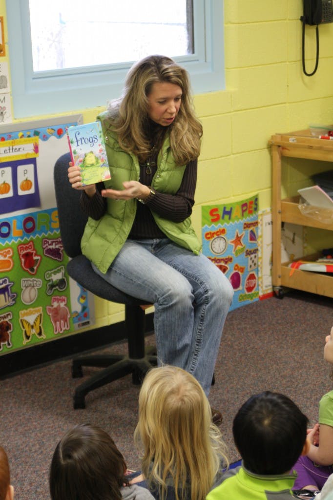 Person sitting on a chair holding a book titled "Frogs" to a group of children