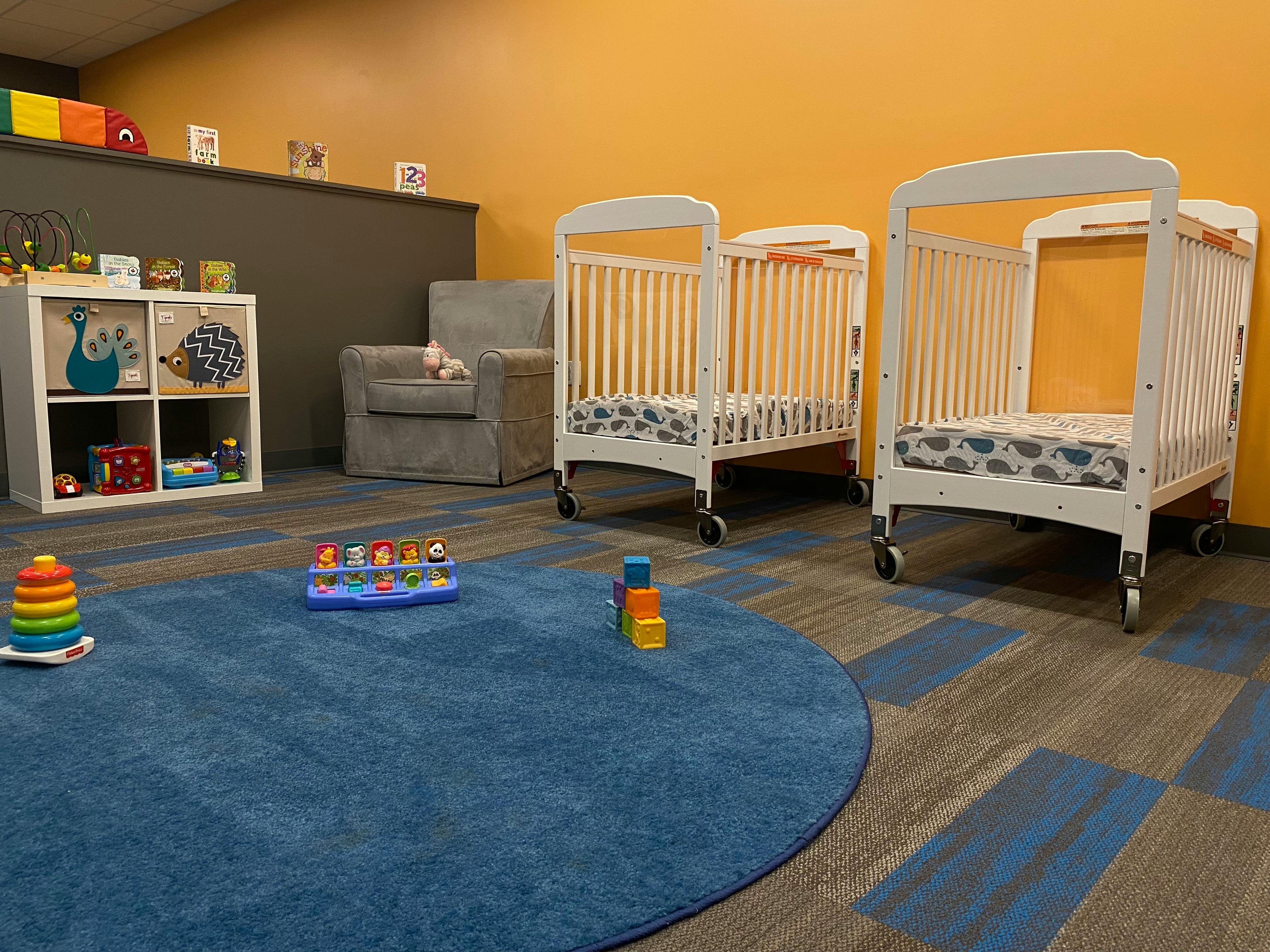 Two cribs in a room with a blue rug and toys