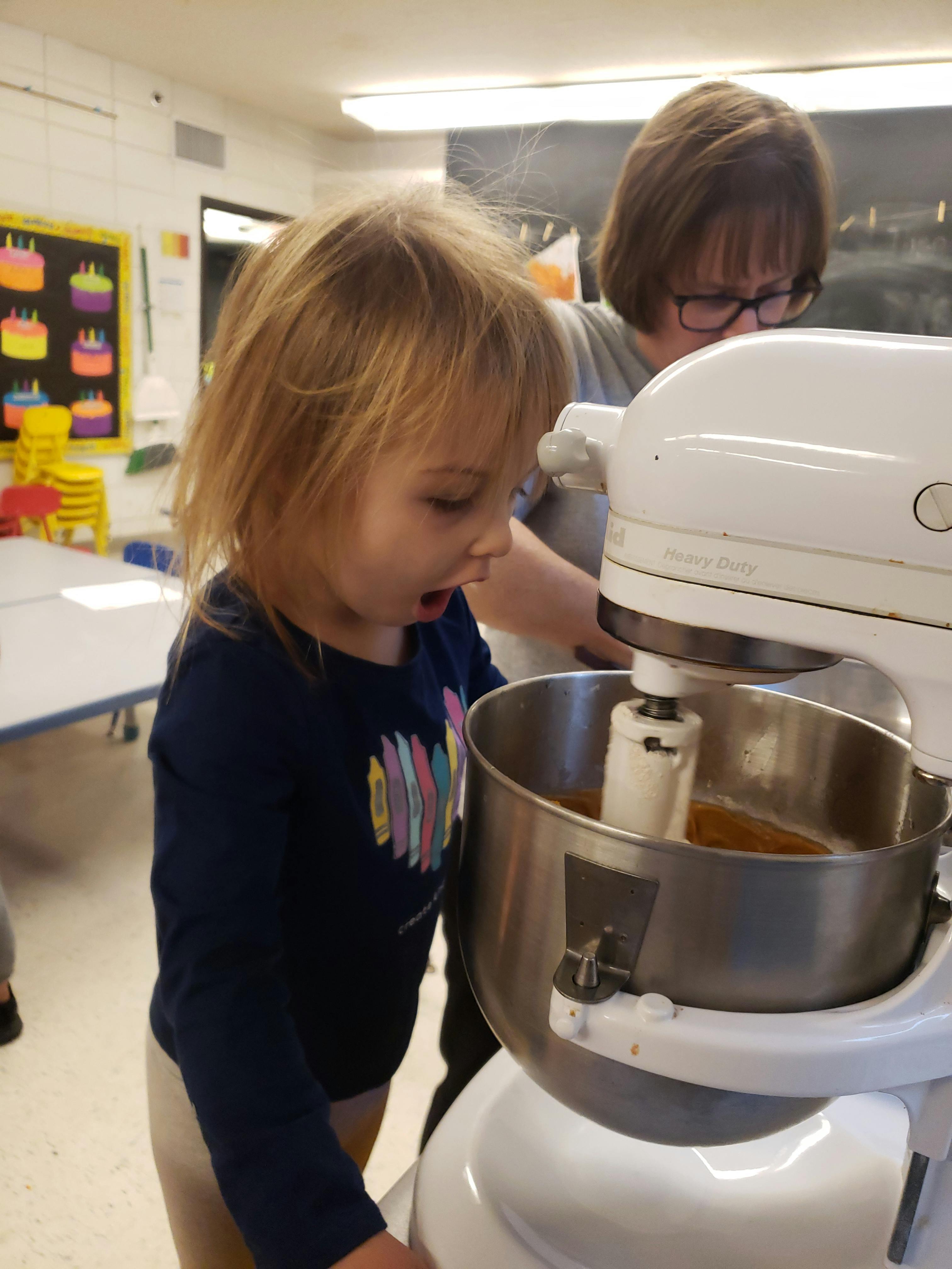 Child looking into a stand mixer with a person in the background
