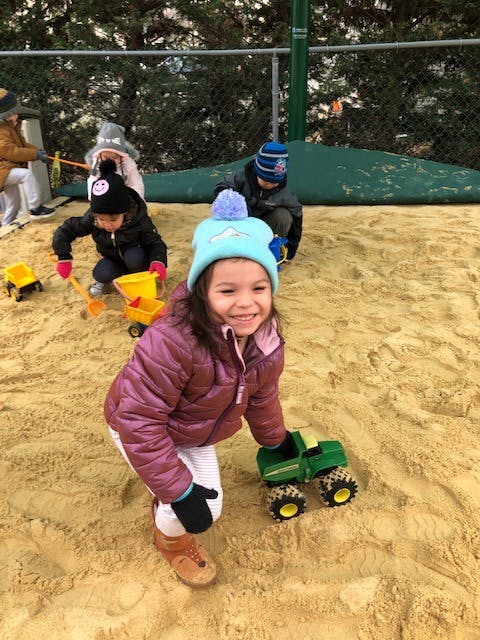 Child in a blue hat playing with a toy truck in a sandbox with other children