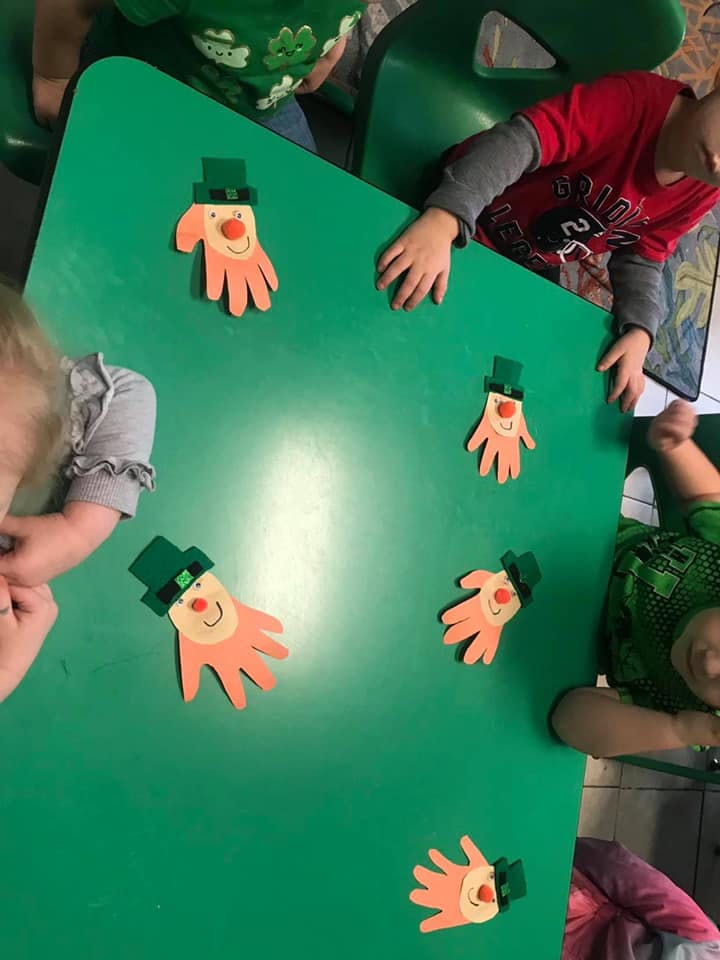 Children around a green table with handprint crafts wearing green hats