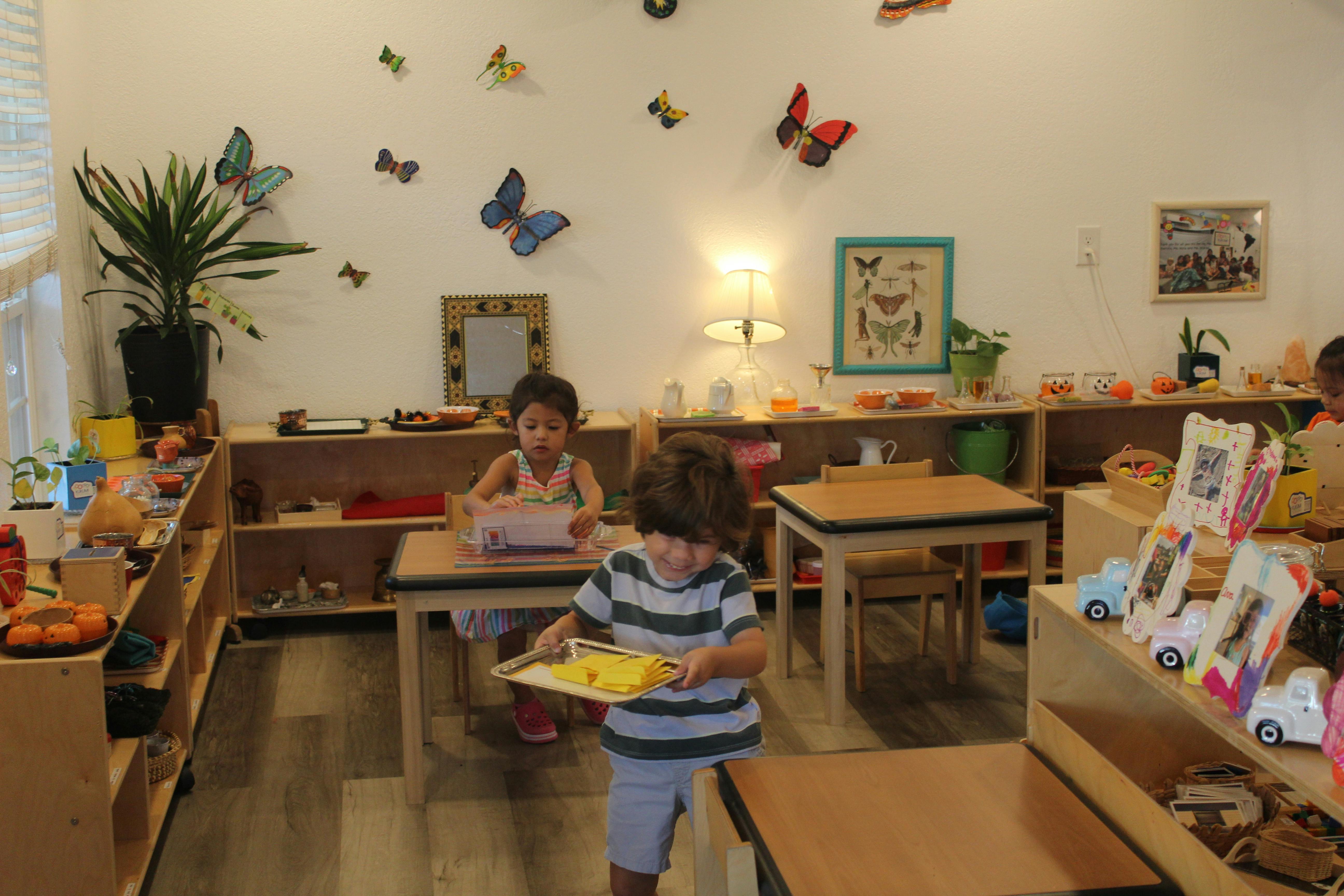 Child carrying a tray in a classroom with another child at a table