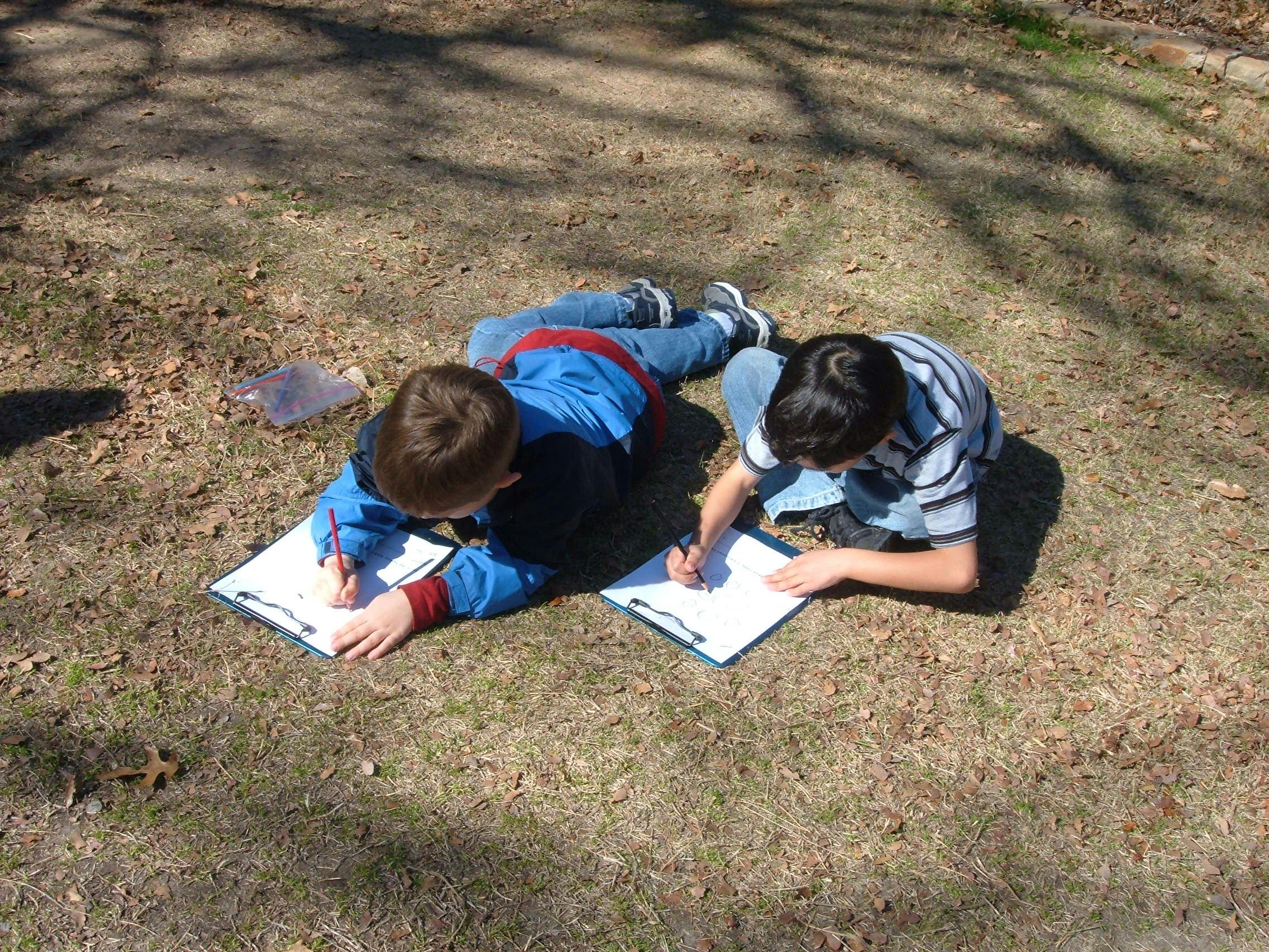 Two children lying on the ground writing on clipboards