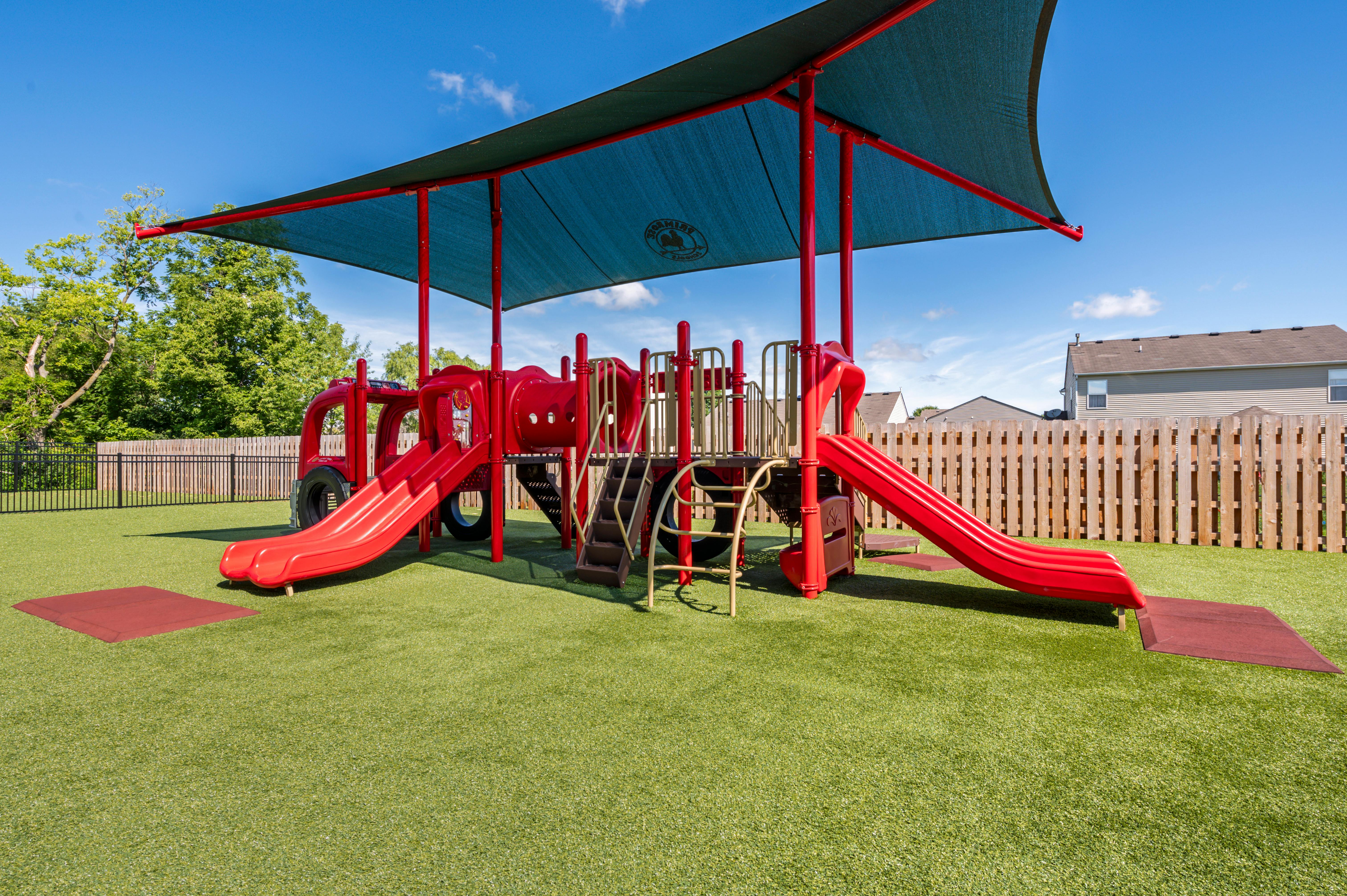 A red playground with slides and climbing elements on green artificial turf under a blue shade canopy