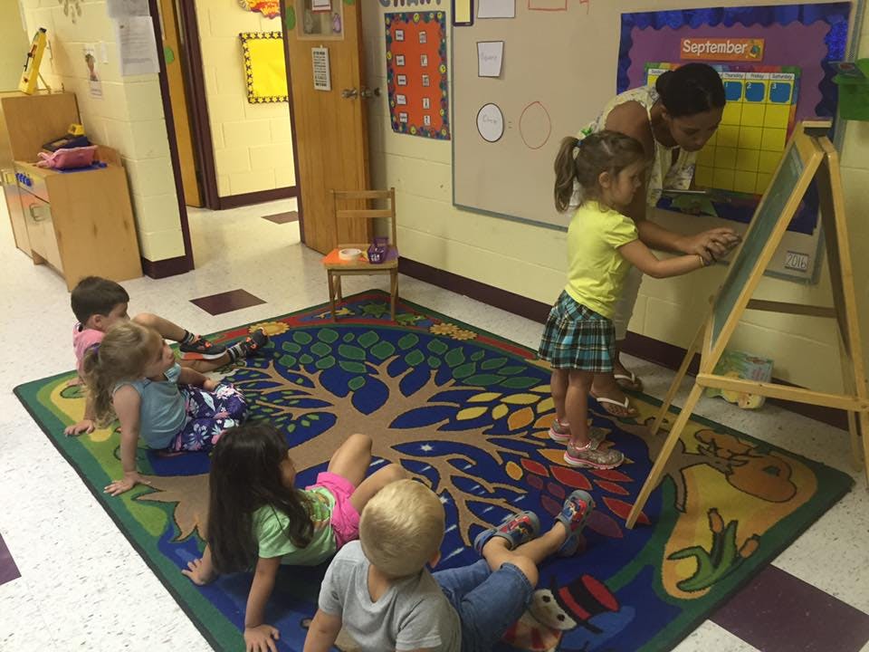 Adult and child at an easel while other children sit on a rug in a classroom