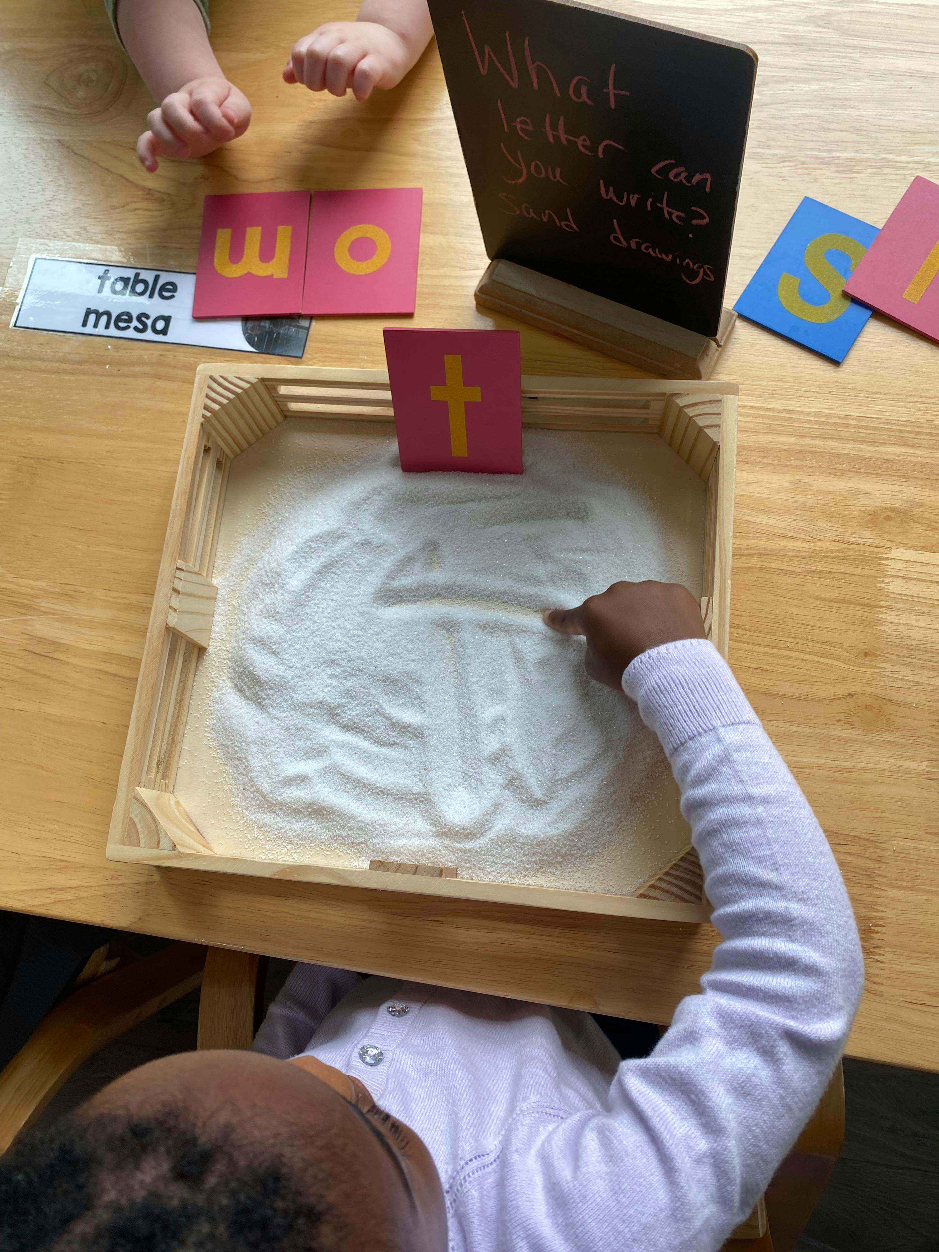 Child tracing a letter in a sand tray with letter cards nearby