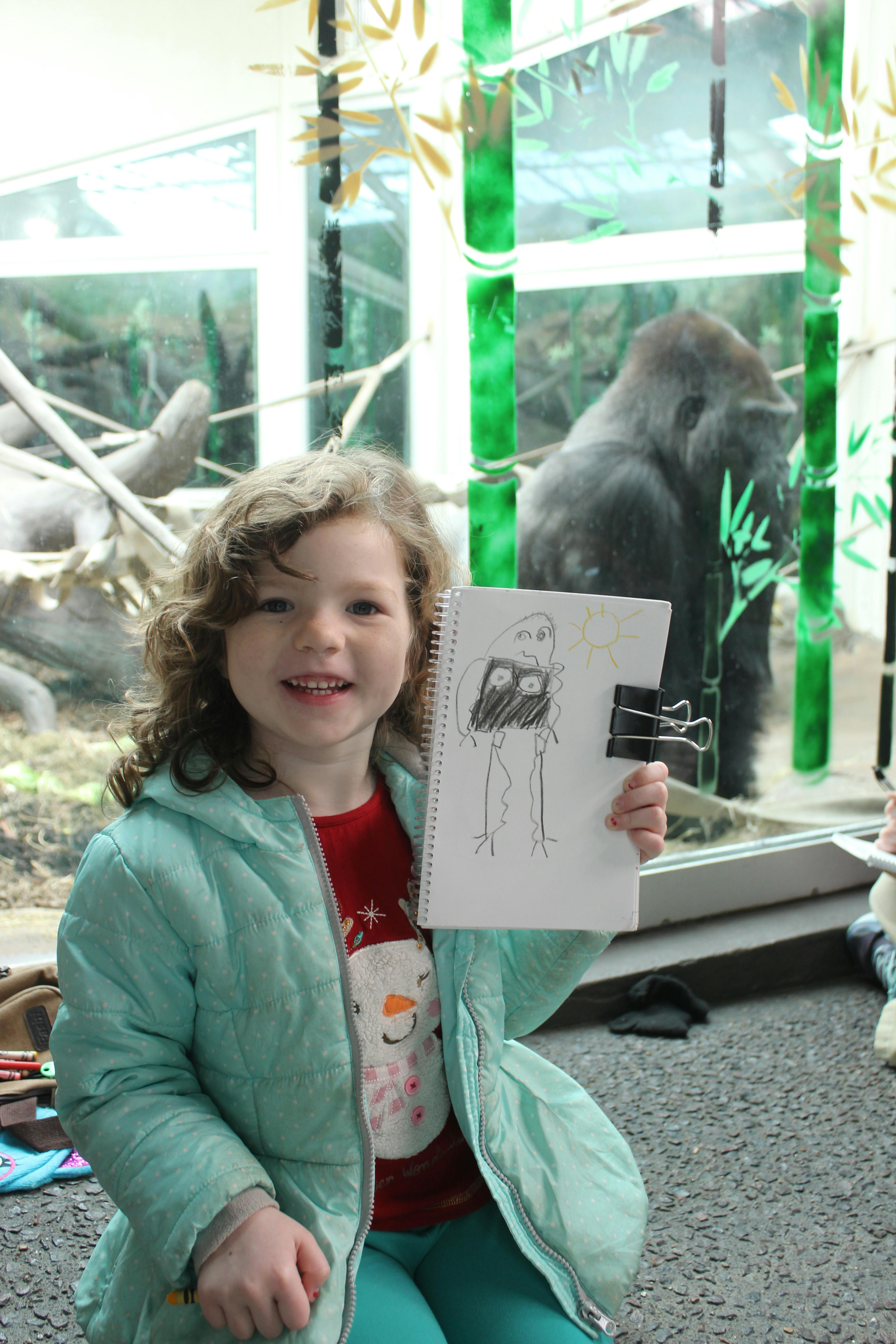 Child holding up a drawing of a gorilla, with a gorilla in the background