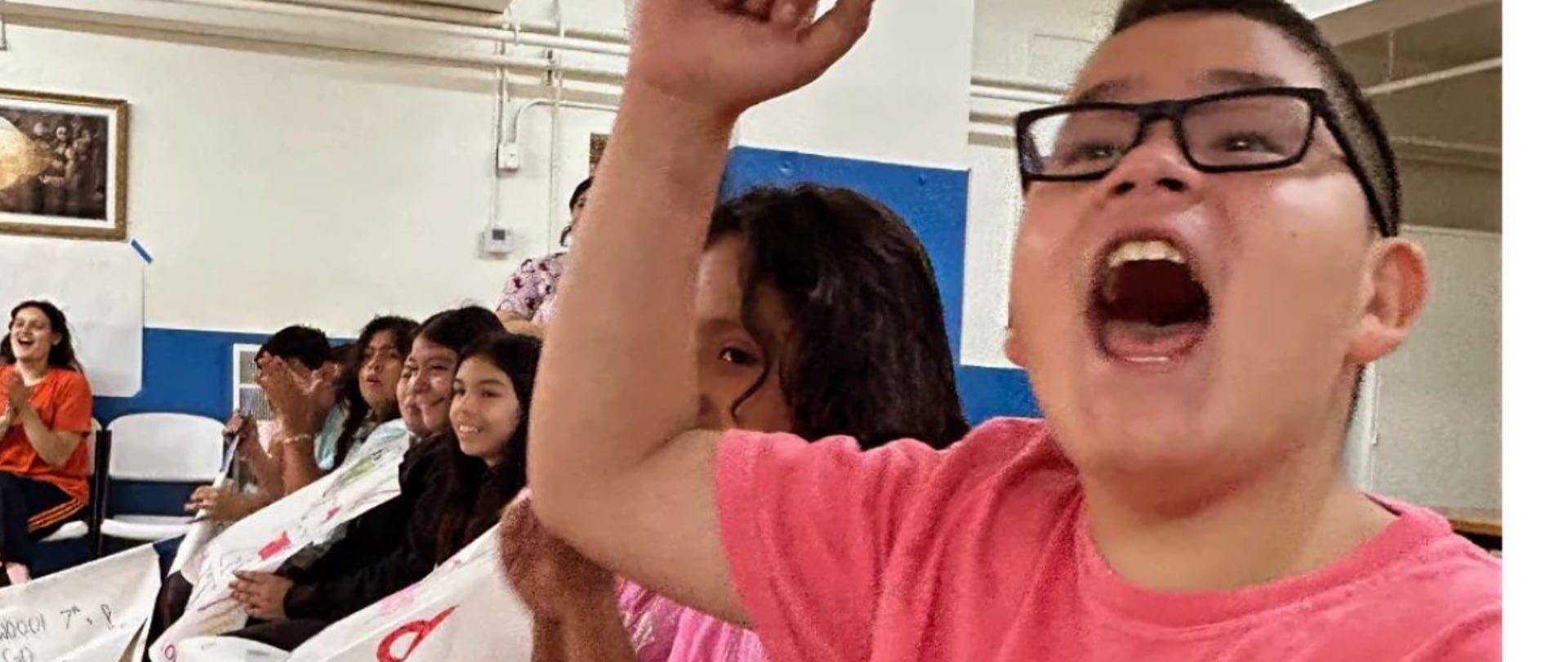 Child in glasses with mouth open and arm raised, people applauding in background