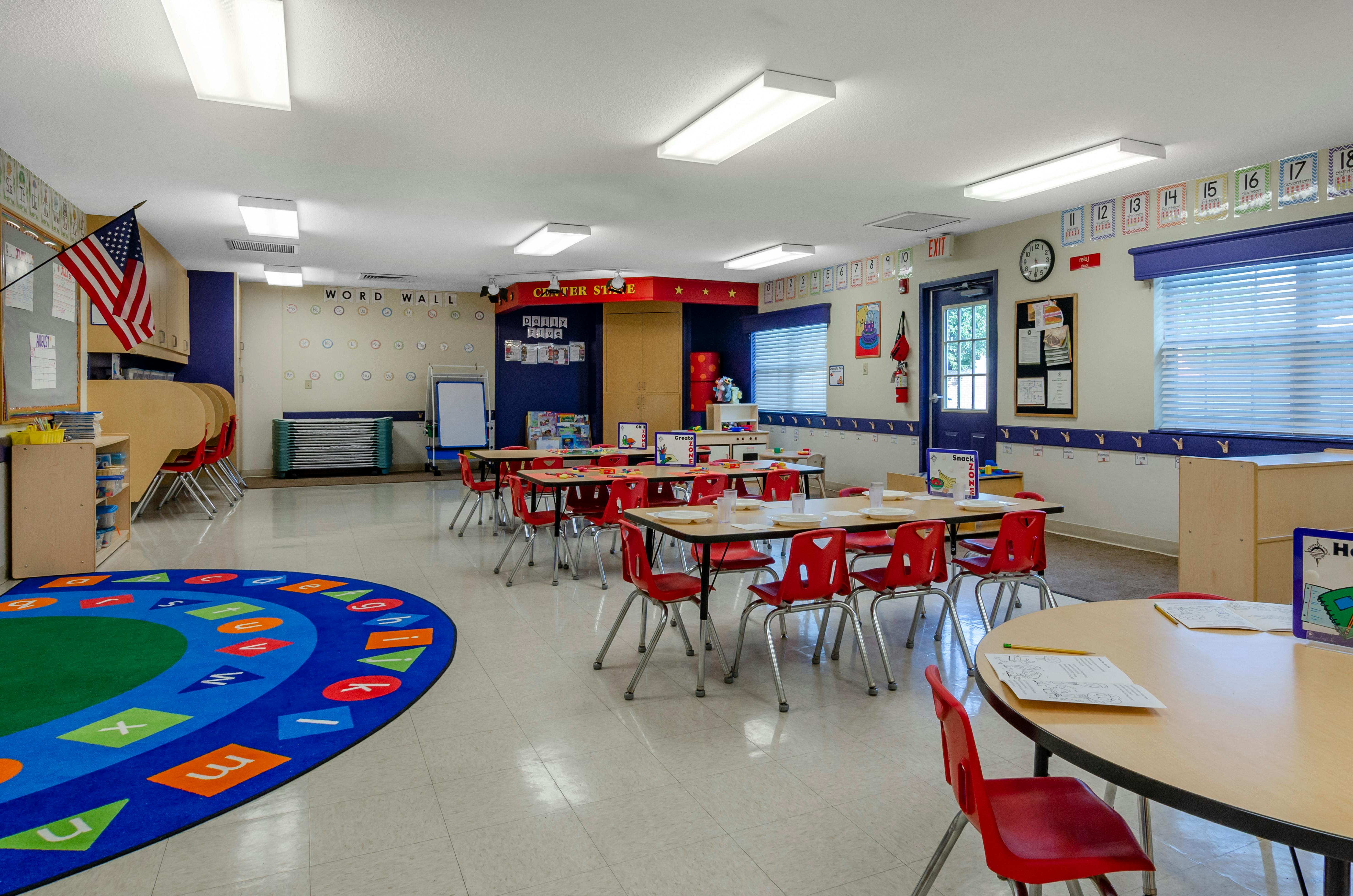 Empty classroom with small tables and red chairs, a colorful rug, and American flag
