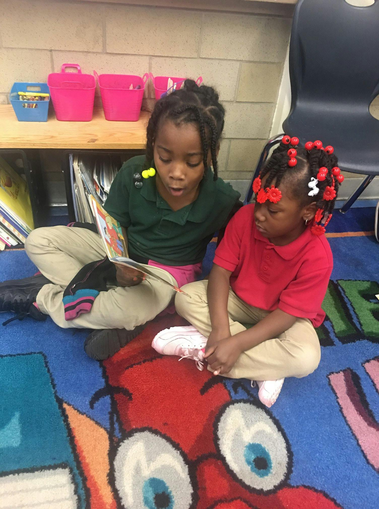 Two children sitting on a rug, one reading a book to the other
