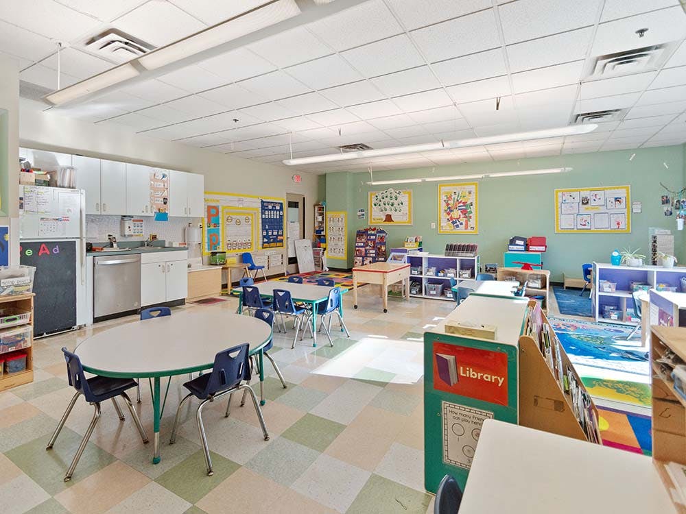 Empty classroom with small tables and chairs, shelves with toys and books