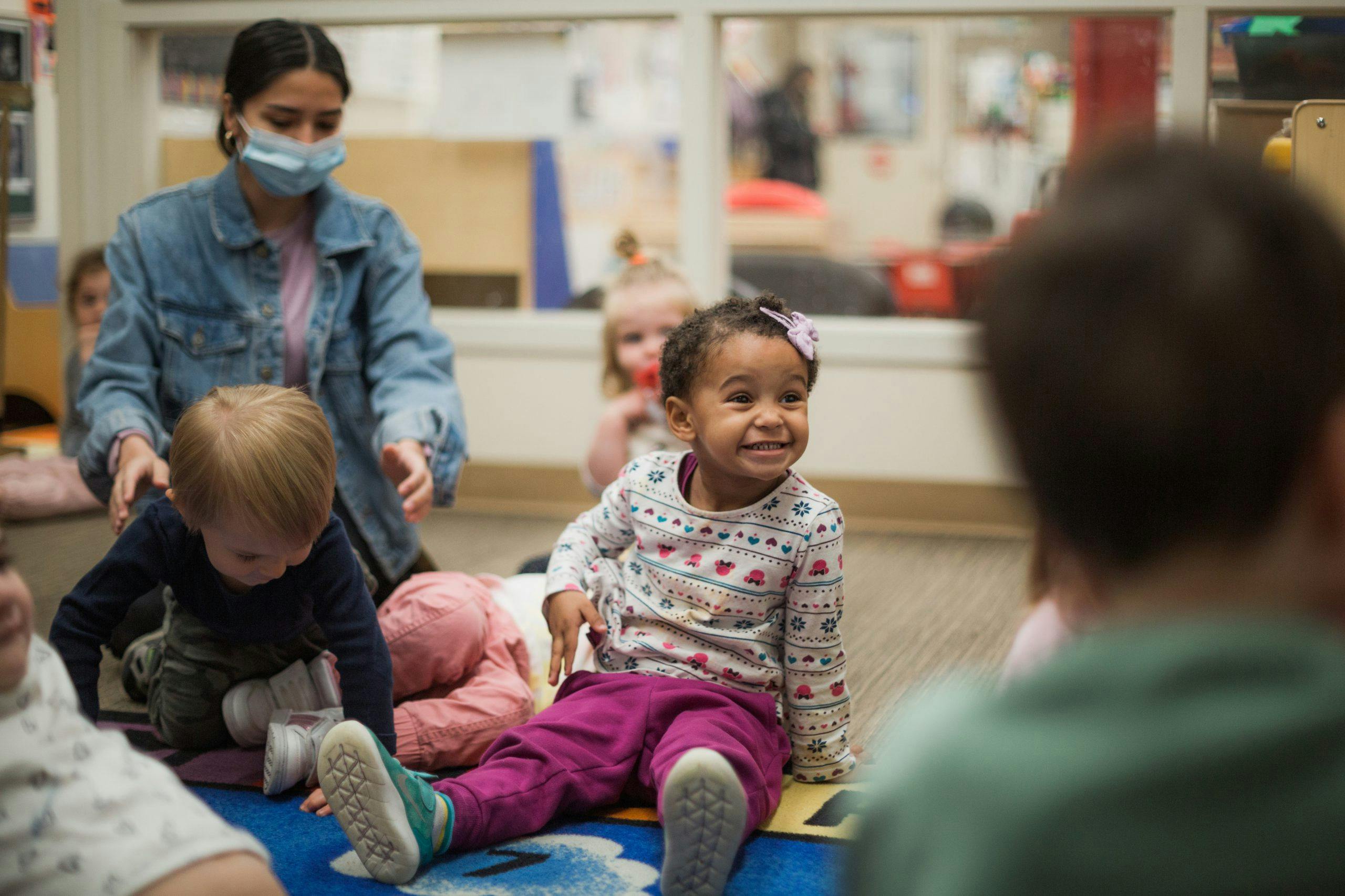 A person in a mask with several young children sitting on a rug in a classroom