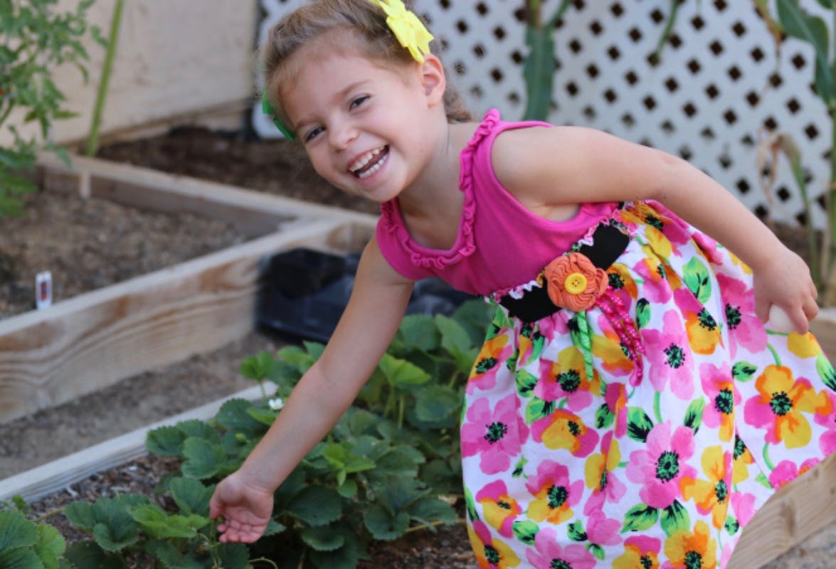 Child in a floral dress reaching towards plants in a garden bed