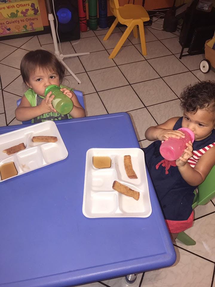 Two children drinking from bottles at a table with food trays