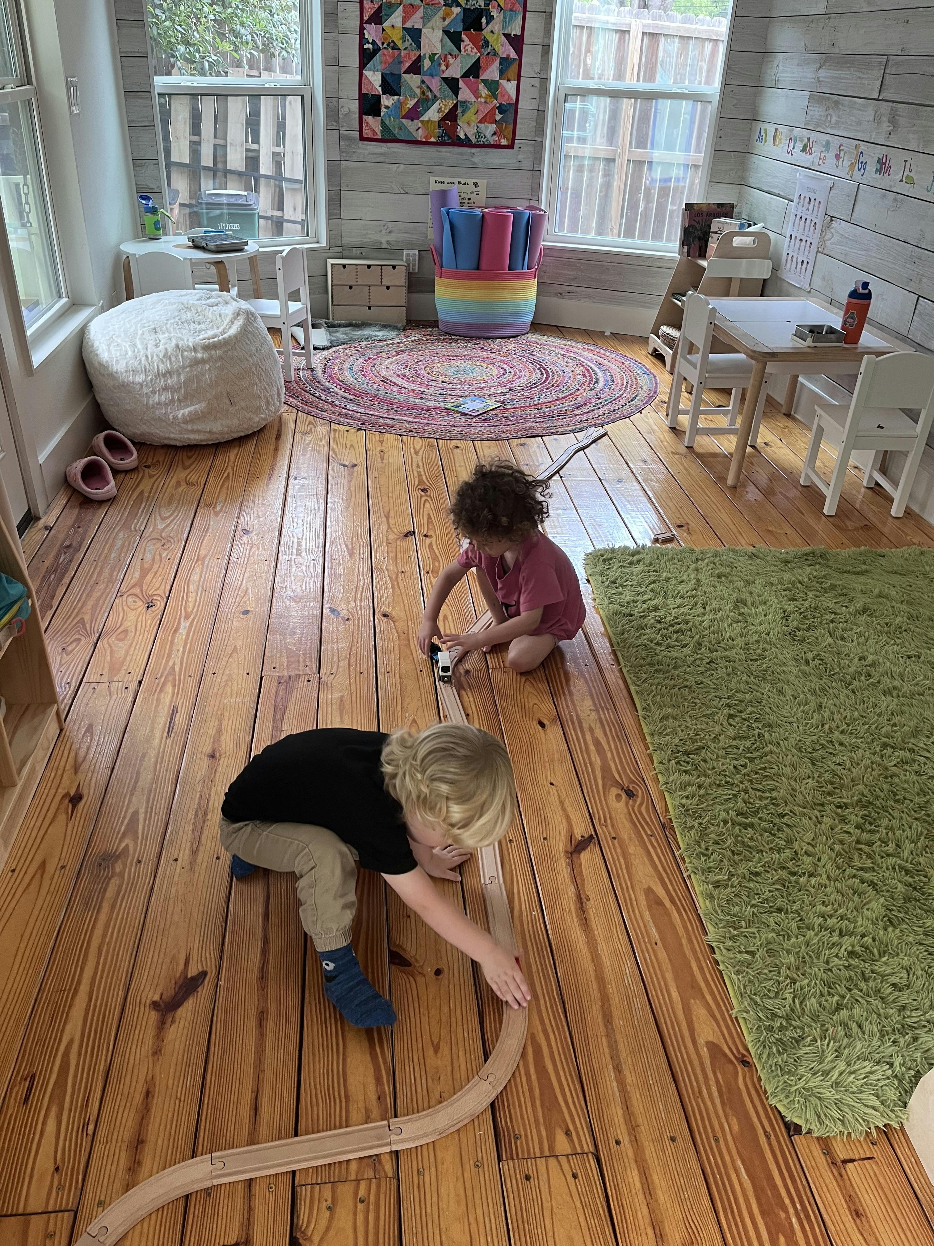 Two children playing with a wooden train track on a wooden floor
