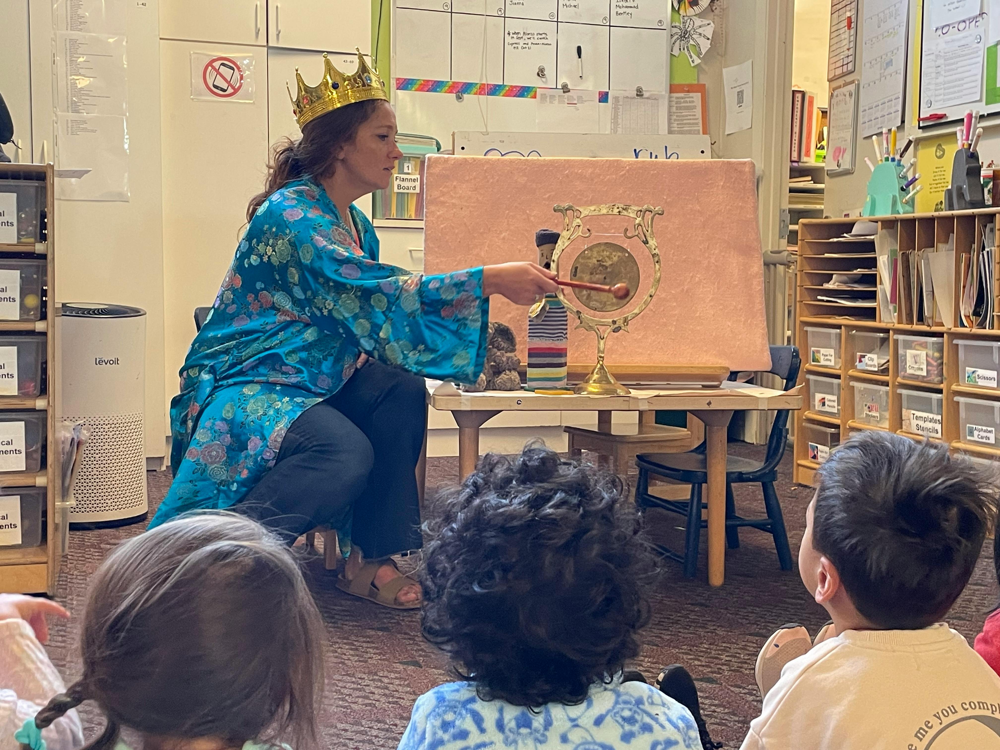 Person in crown playing a gong for children in a classroom