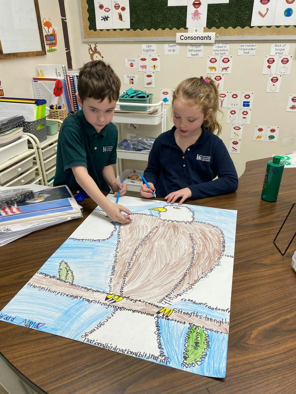 Two children coloring a large drawing of a bald eagle in a classroom