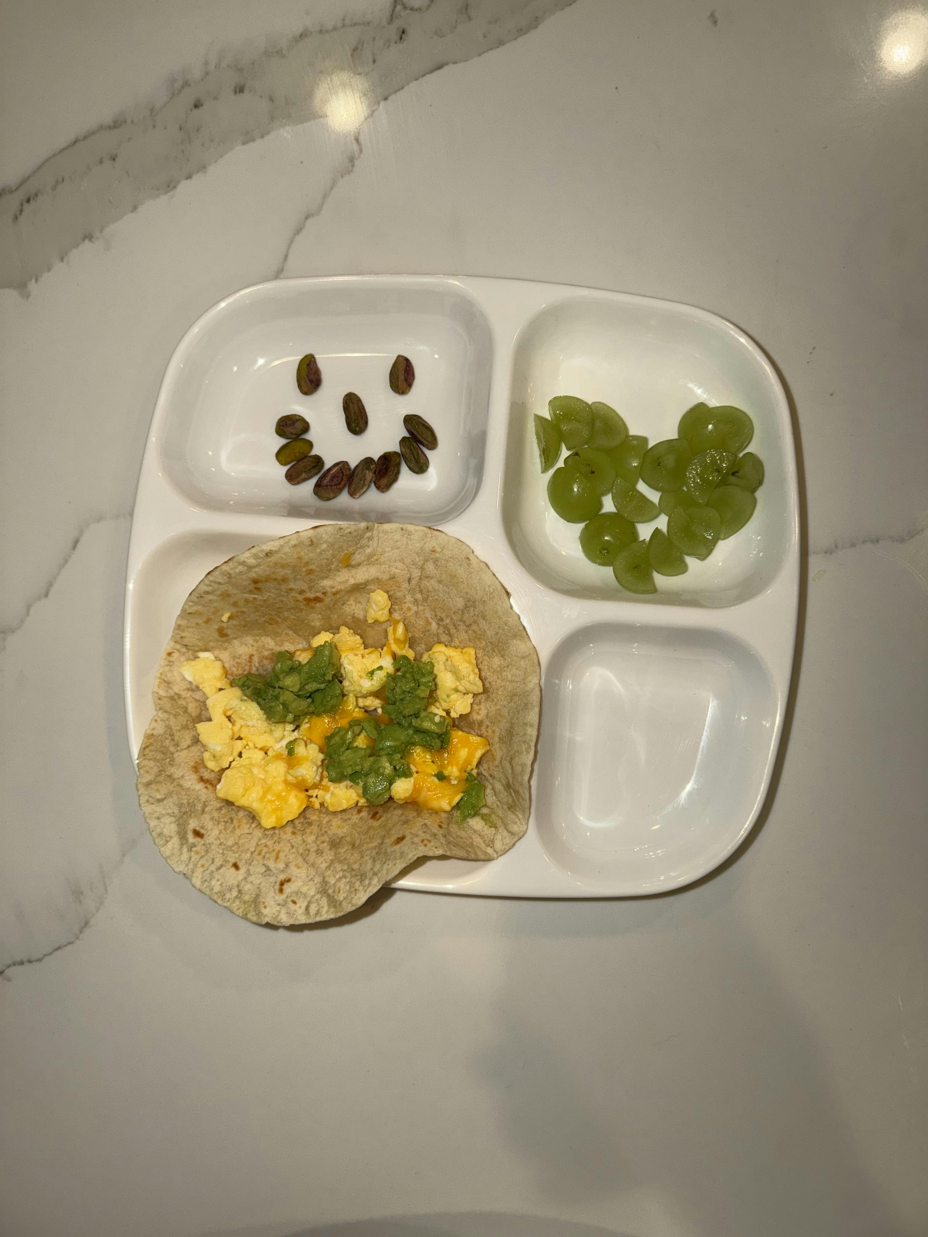 Sectioned plate with scrambled eggs on a tortilla, grapes, and raisins arranged in a smile