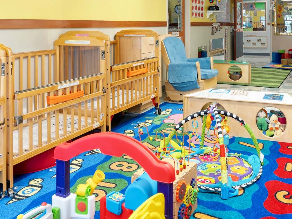 Cribs, a blue rocking chair, and play mats on a colorful rug in a daycare room