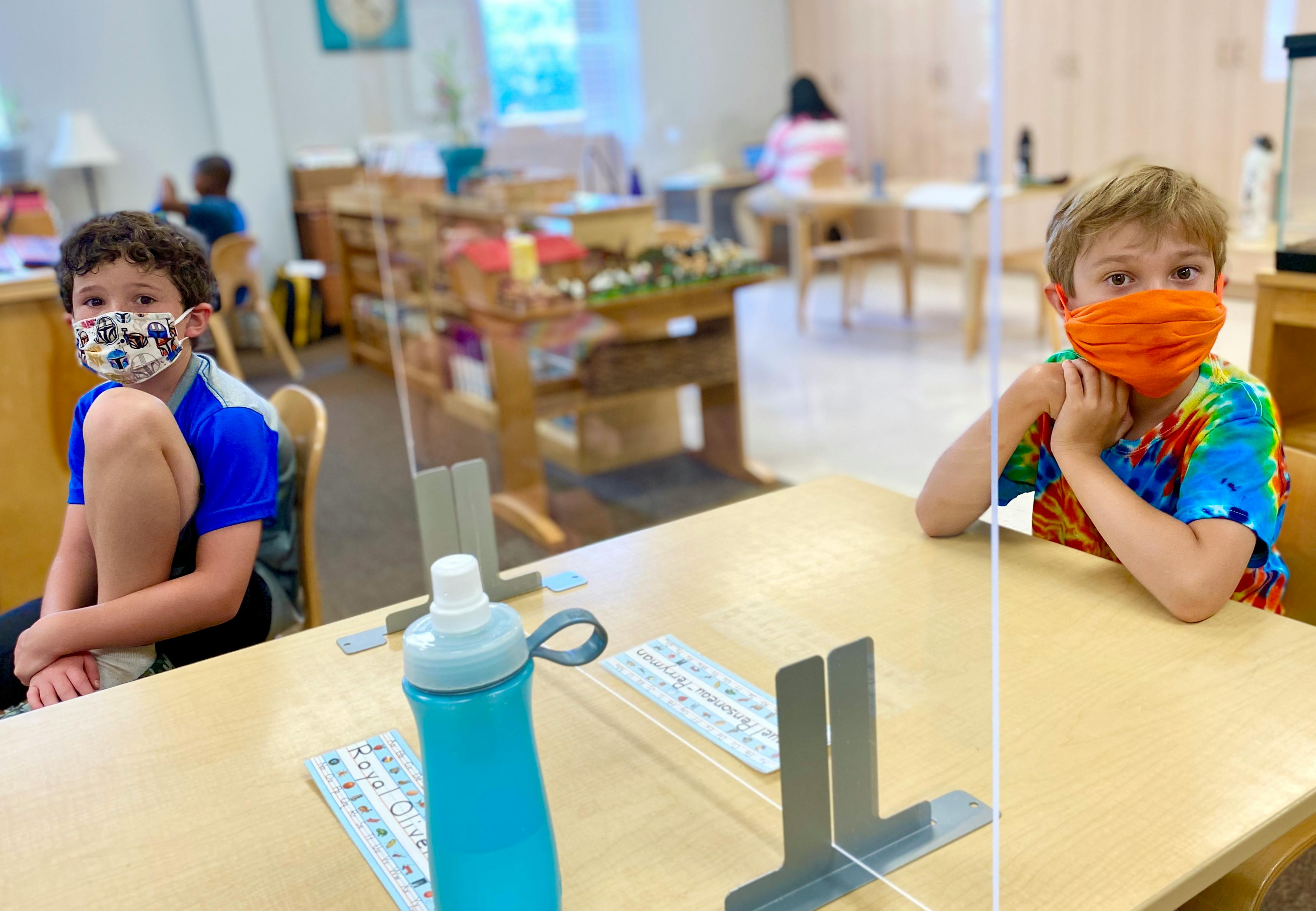 Two children wearing face masks sitting at desks with clear dividers