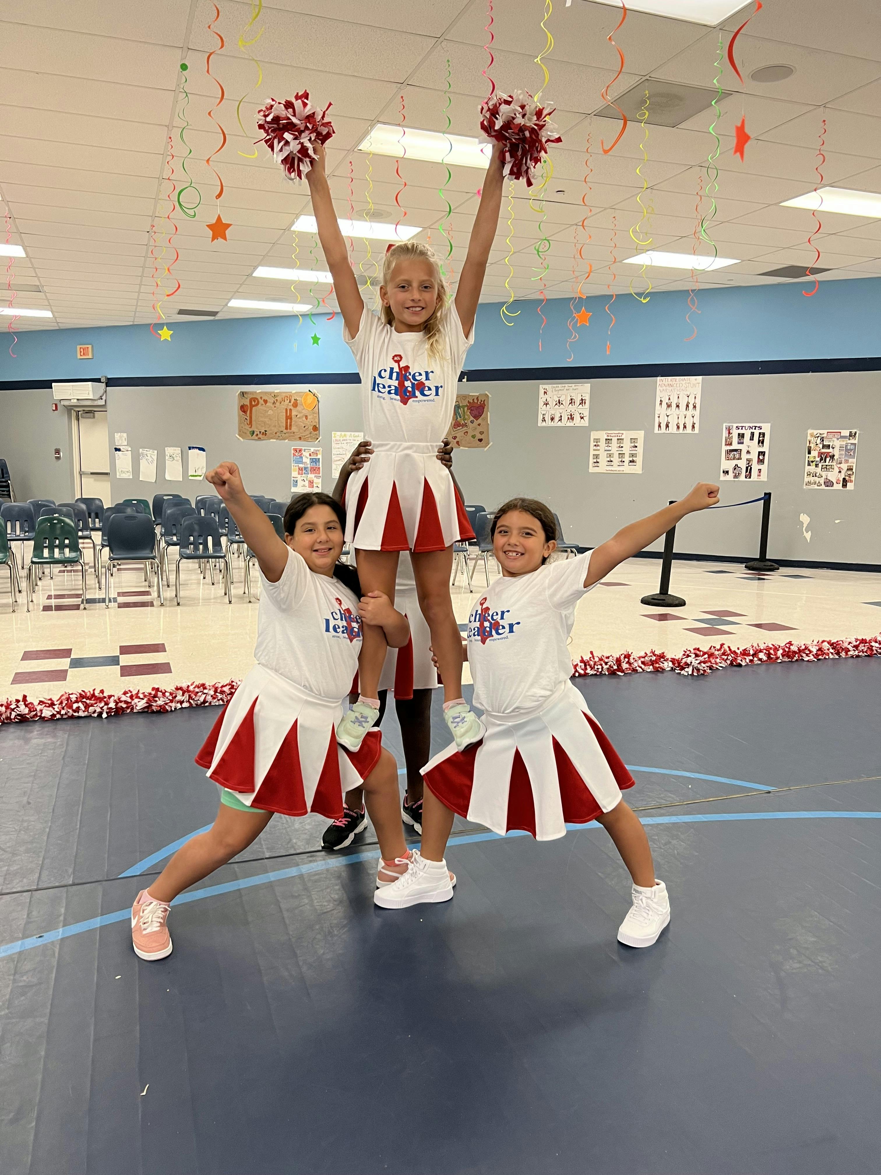 Three children in cheerleader outfits forming a pyramid with pompoms