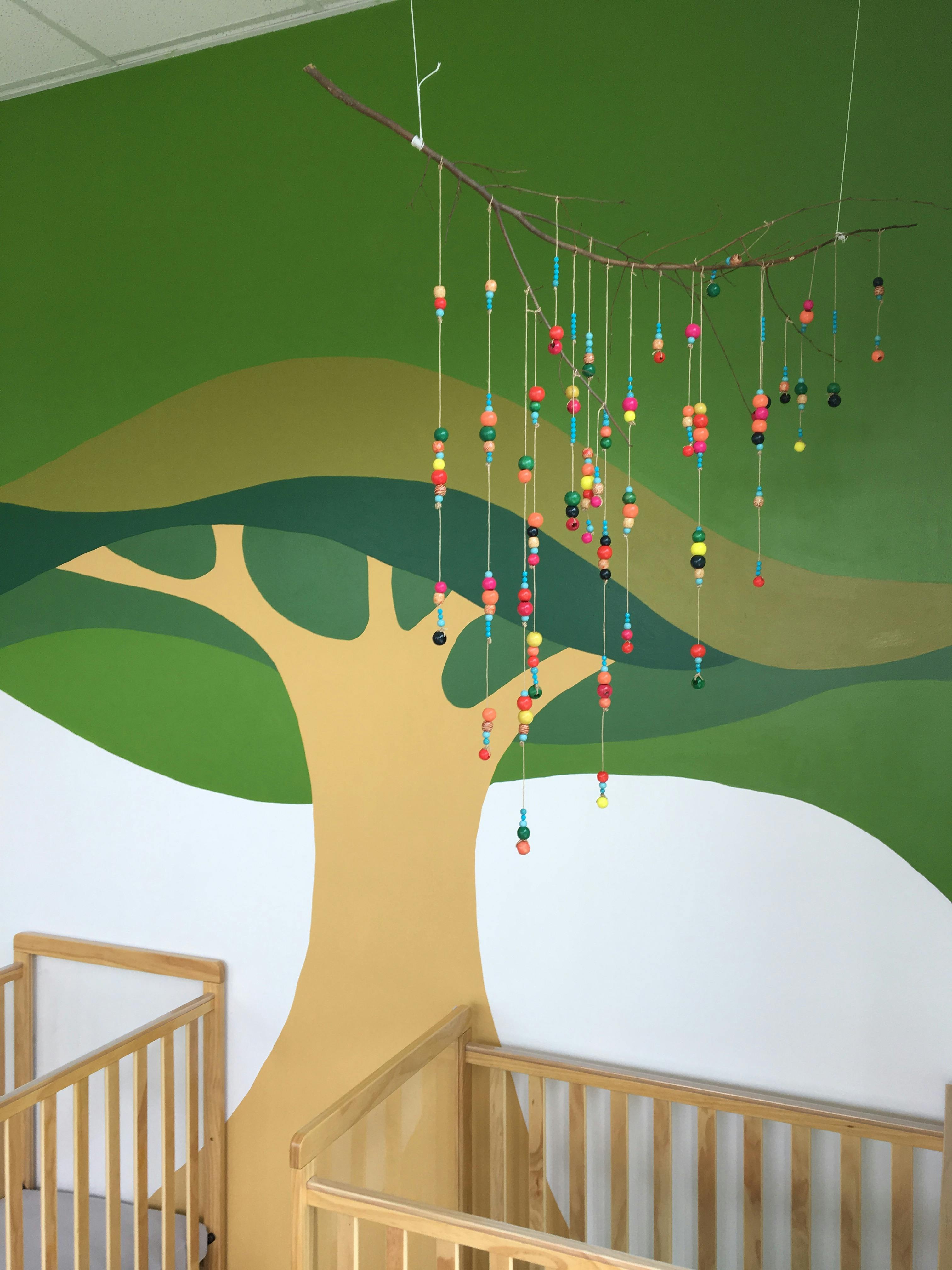 Beaded mobile hanging above cribs with a tree mural on the wall