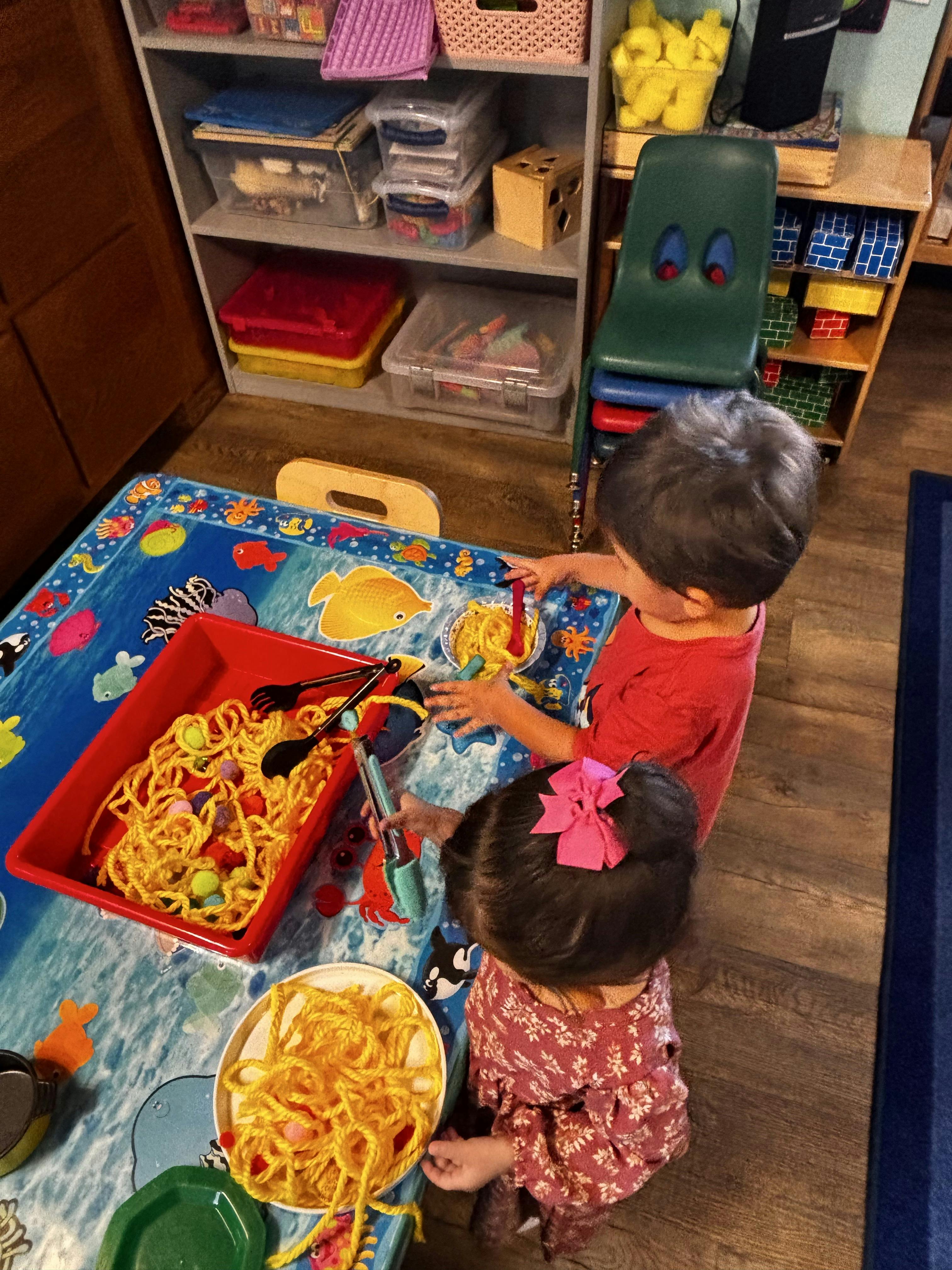 Two children playing with yarn and pom-poms on a table with an ocean mat