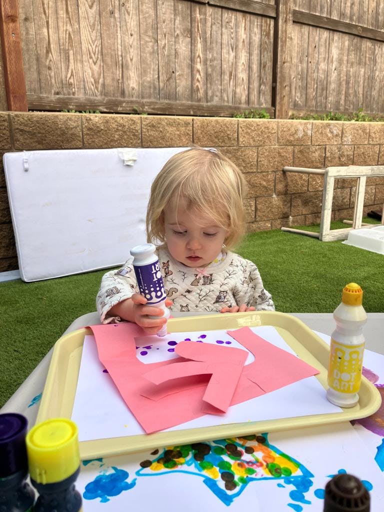 Child holding a bottle of paint over pink paper on a tray
