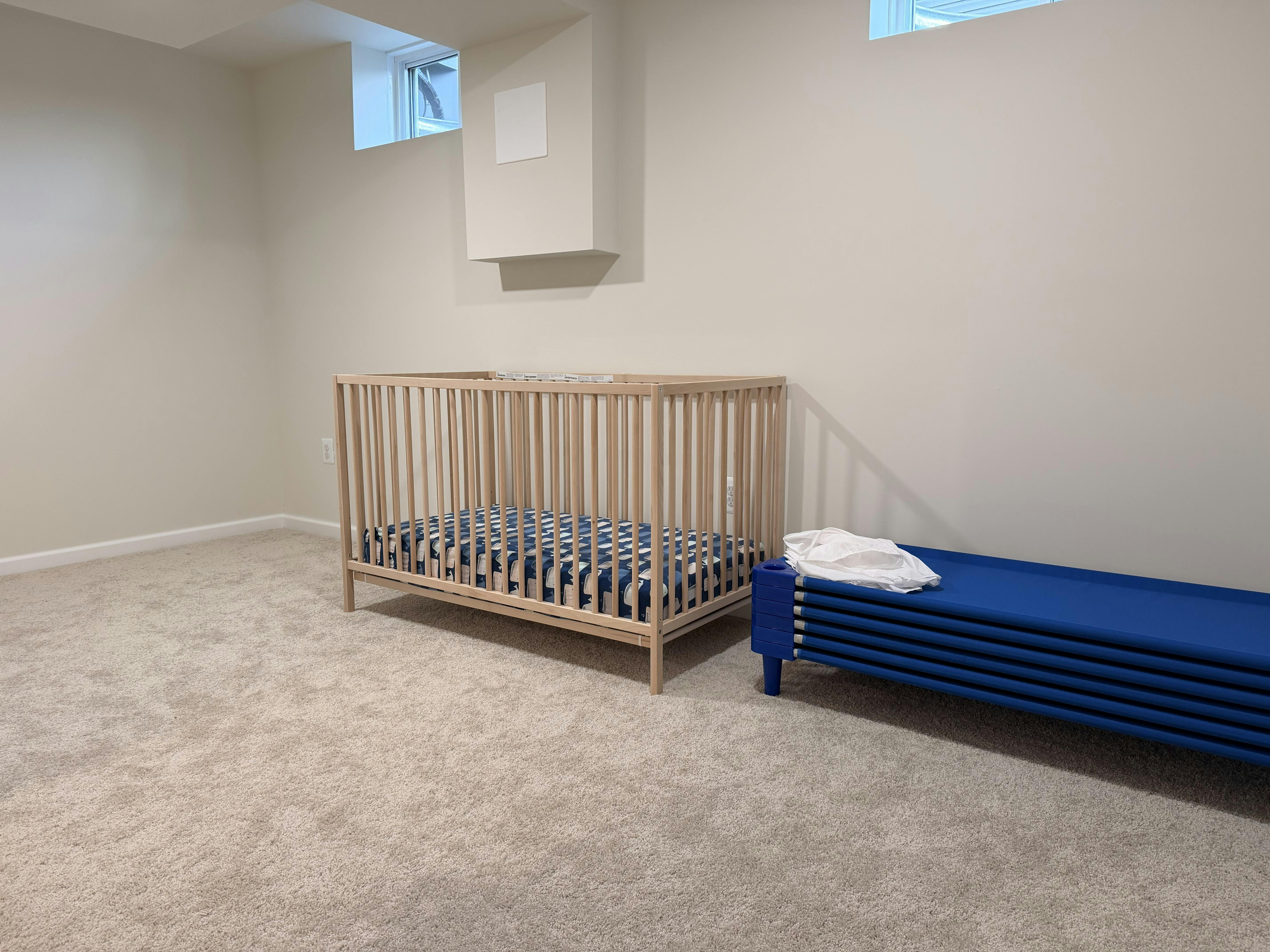 Crib with a patterned mattress and a stack of blue cots in a carpeted room