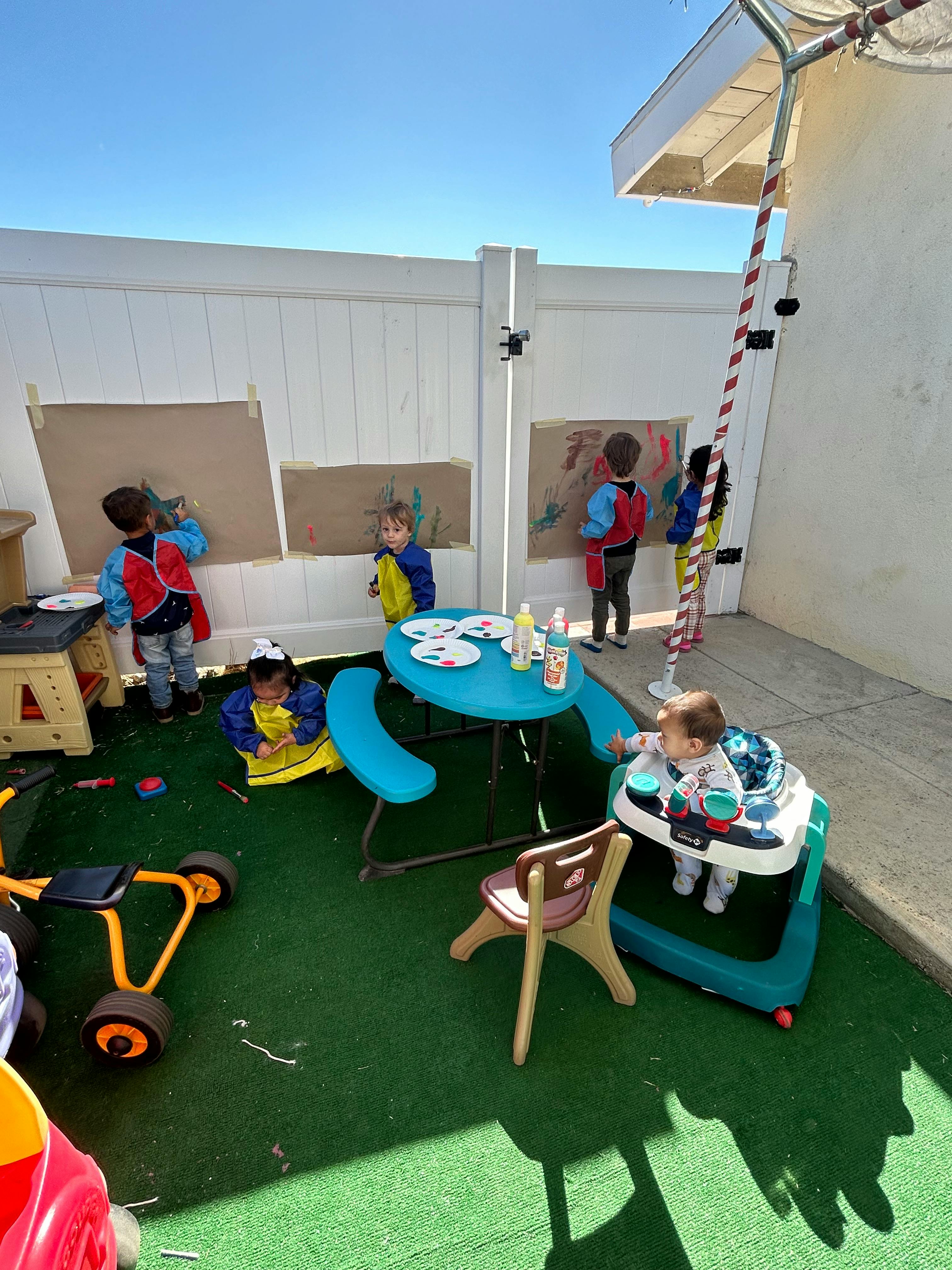 Children painting on paper attached to a white fence outdoors