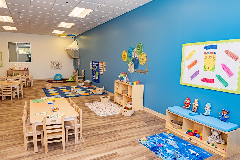 Empty classroom with small tables and chairs, play mats, and shelves with toys and learning materials