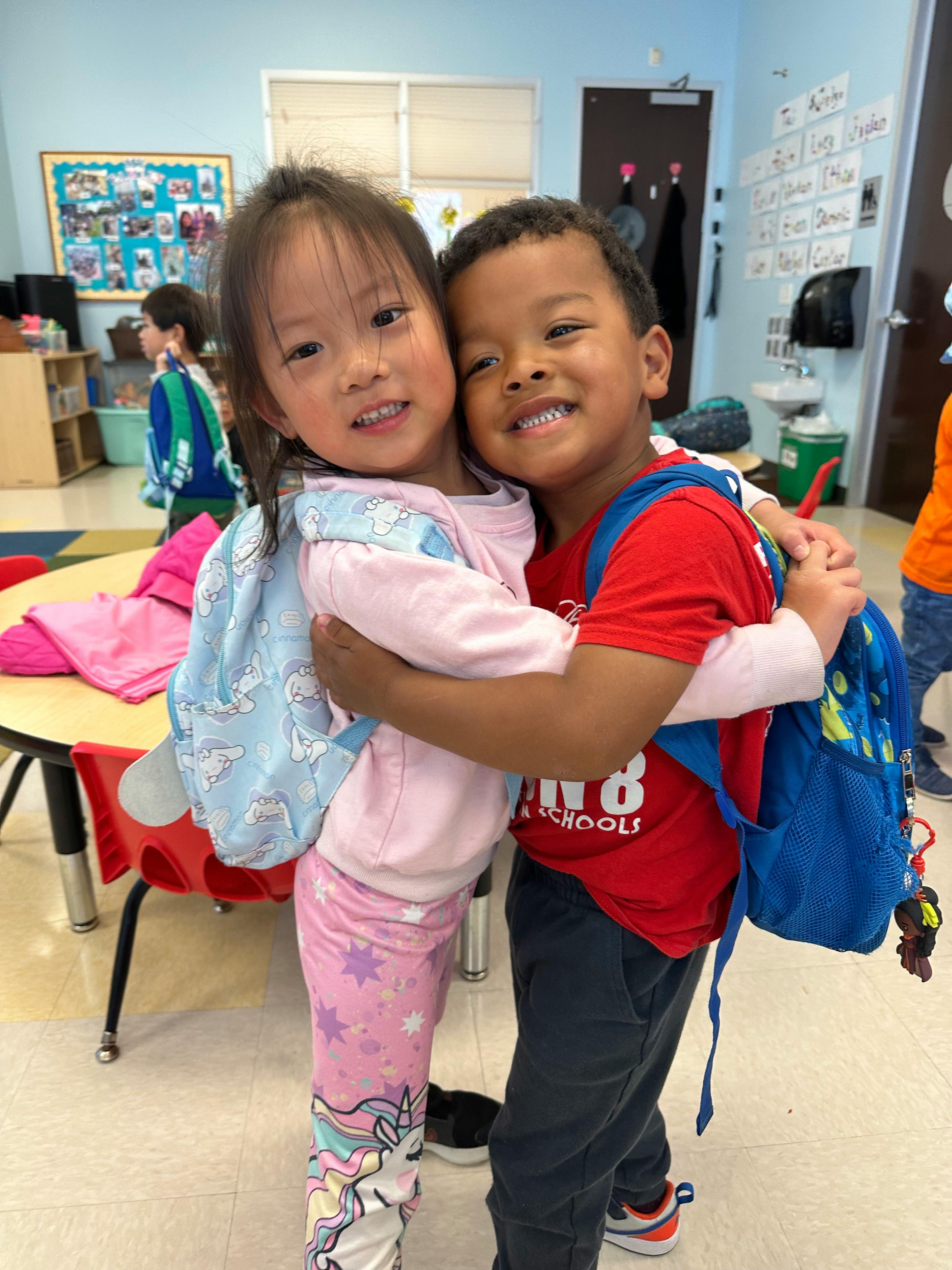 Two children with backpacks hugging in a classroom