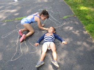 Child drawing chalk outline around another child lying on pavement