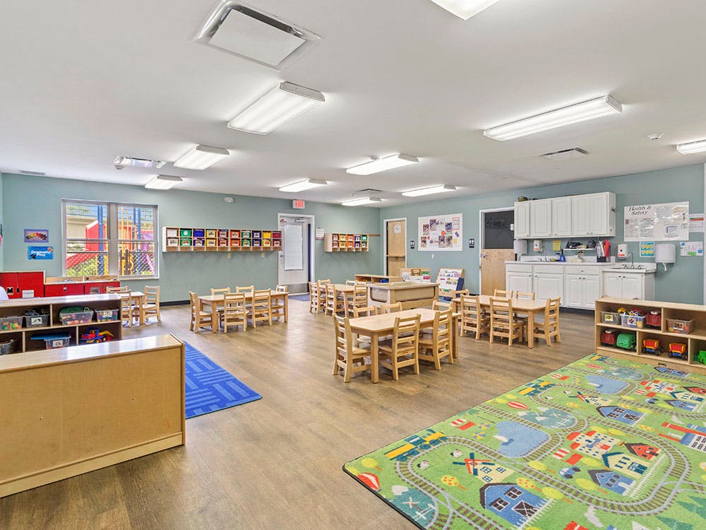 Classroom with small wooden tables and chairs, and a play mat with a road design