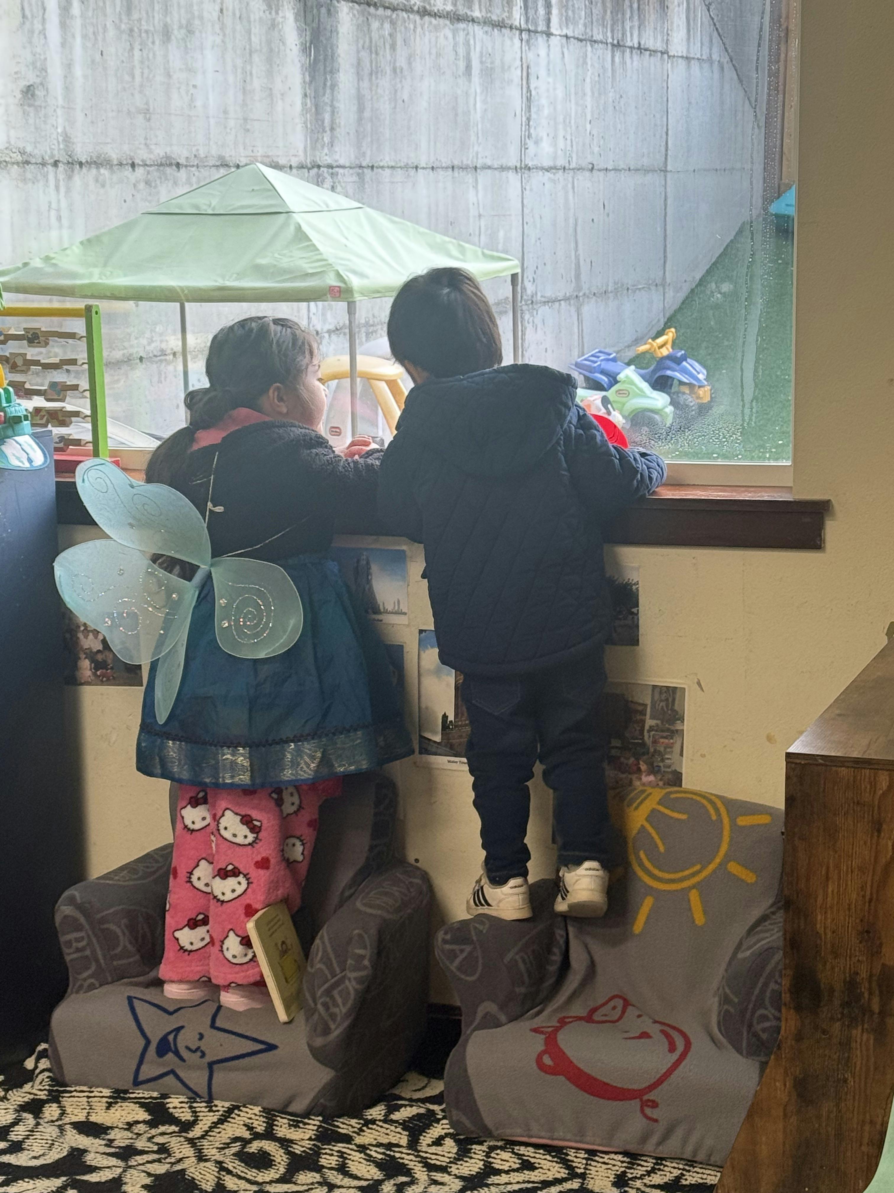 Two children standing on chairs and looking out a window