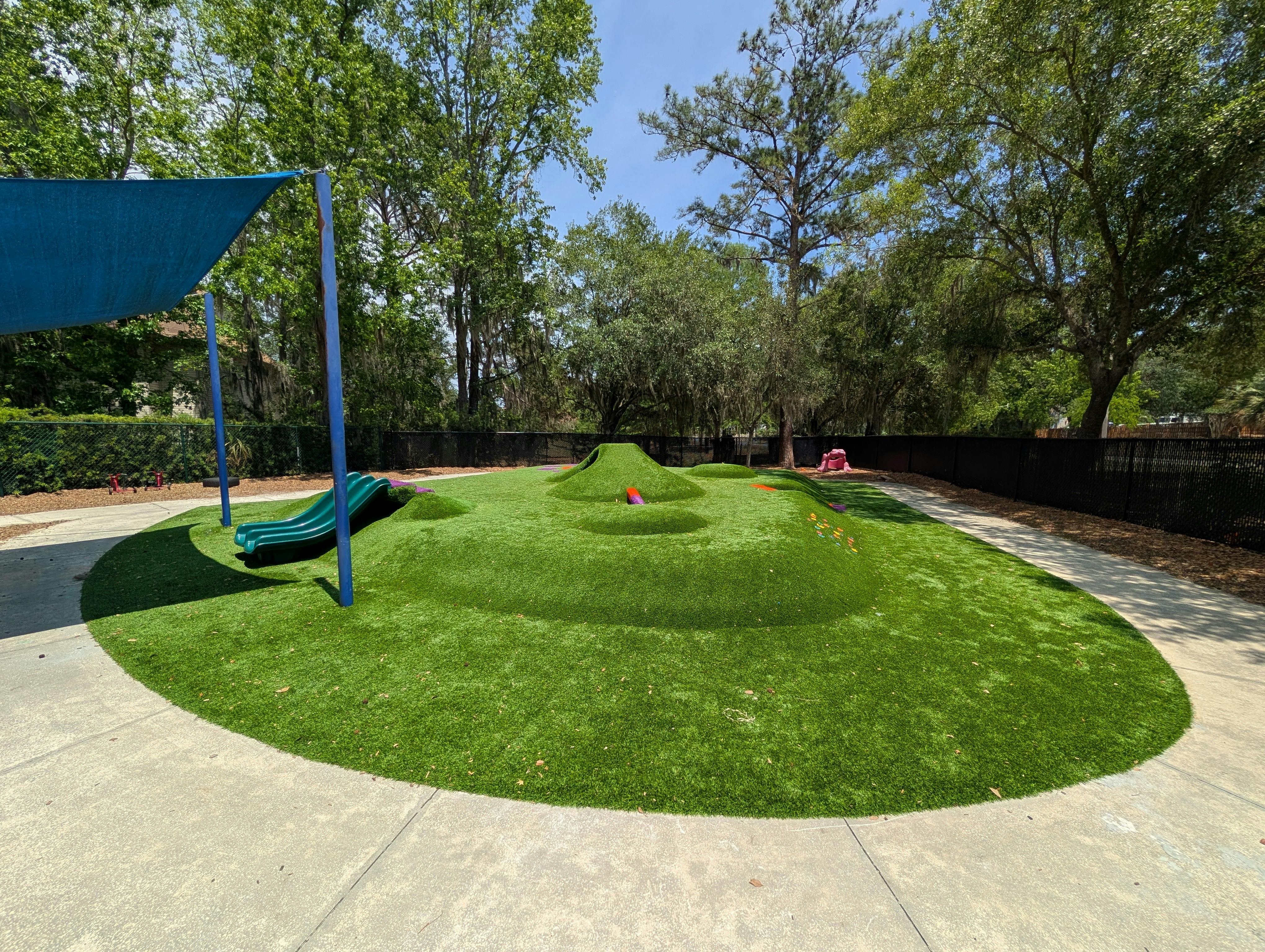 Playground with green turf mounds and slides, bordered by a concrete path and trees