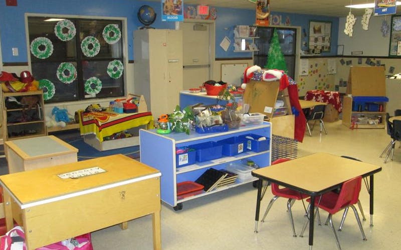 An empty classroom with small tables and chairs, shelves, and a Christmas tree