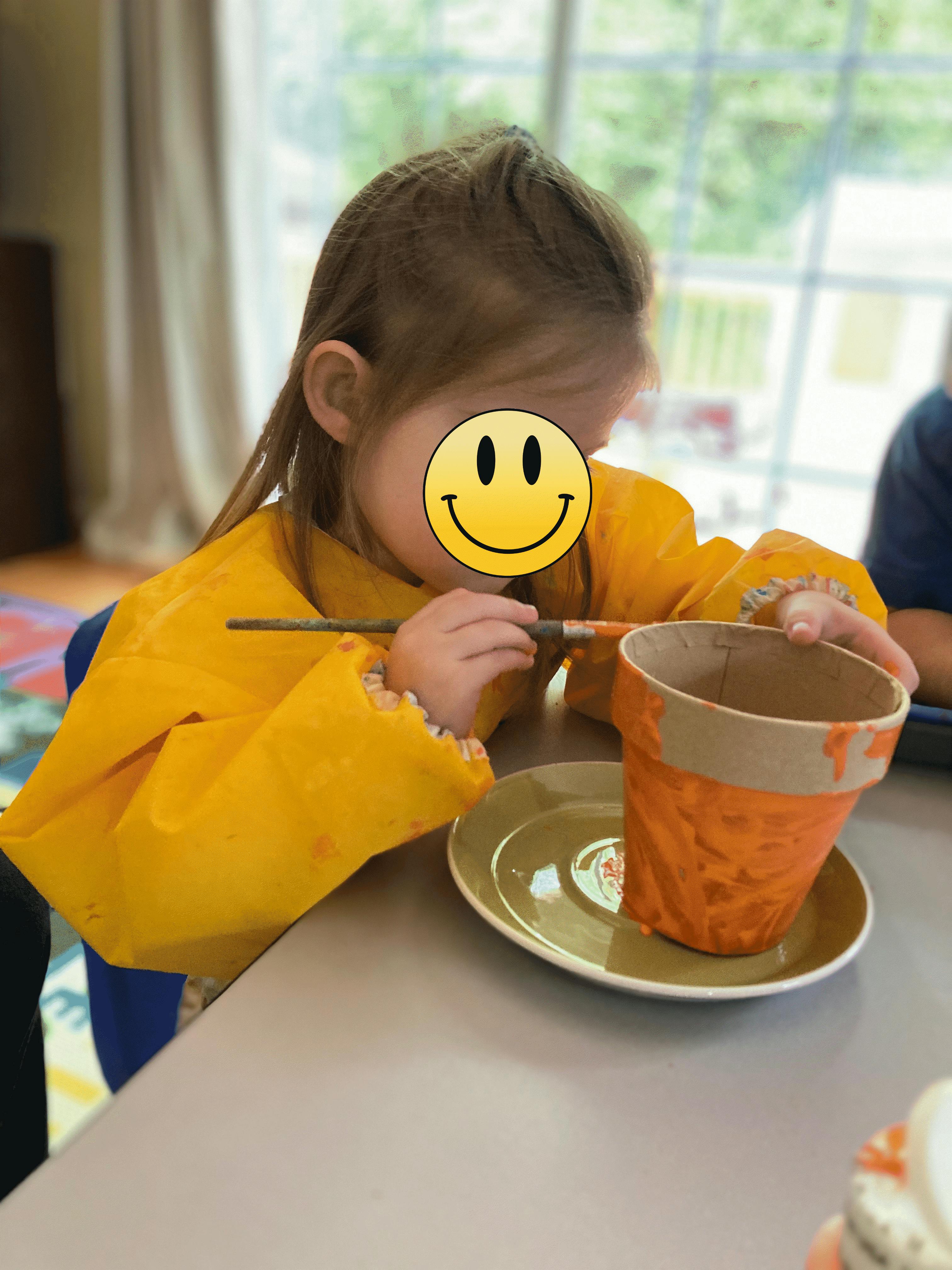 Child wearing a yellow apron painting a pot at a table