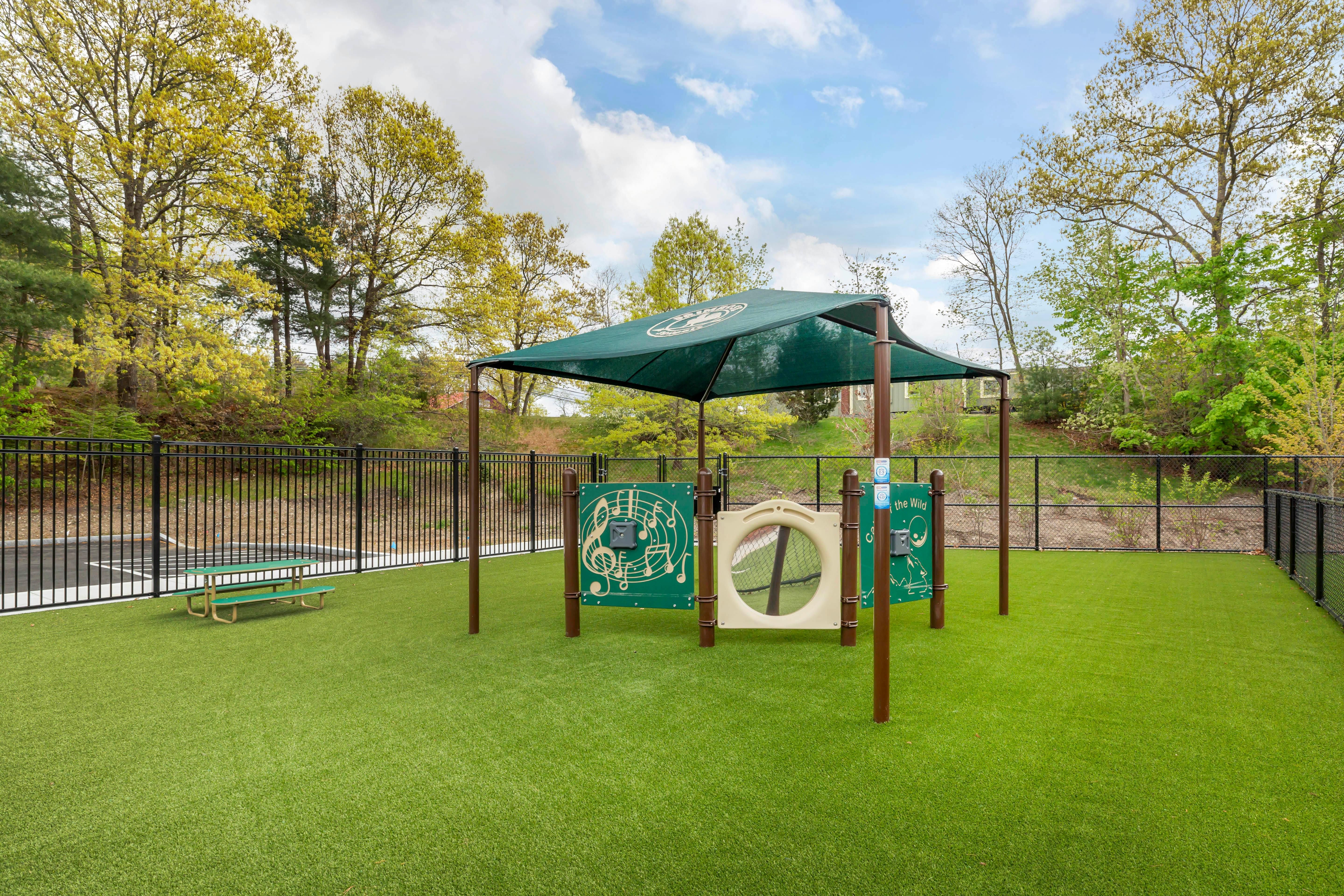 A small green outdoor playset with a shade canopy on artificial turf, next to a black fence