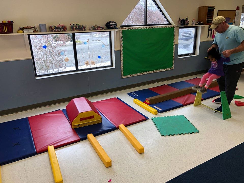 Adult helping a child on a balance beam in a room with gym mats