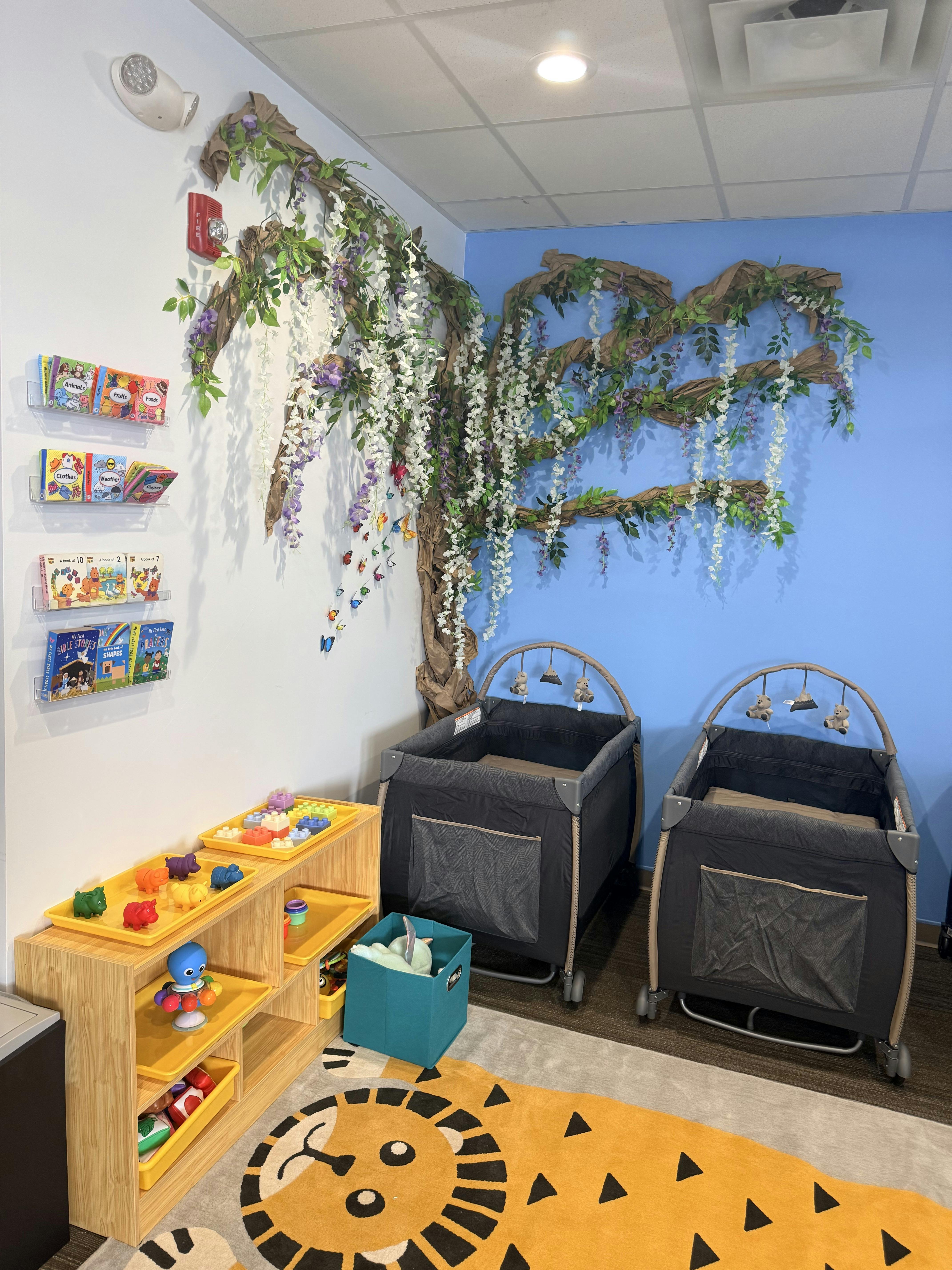 A colorful play area with cribs, shelves of toys and books, and a tree mural