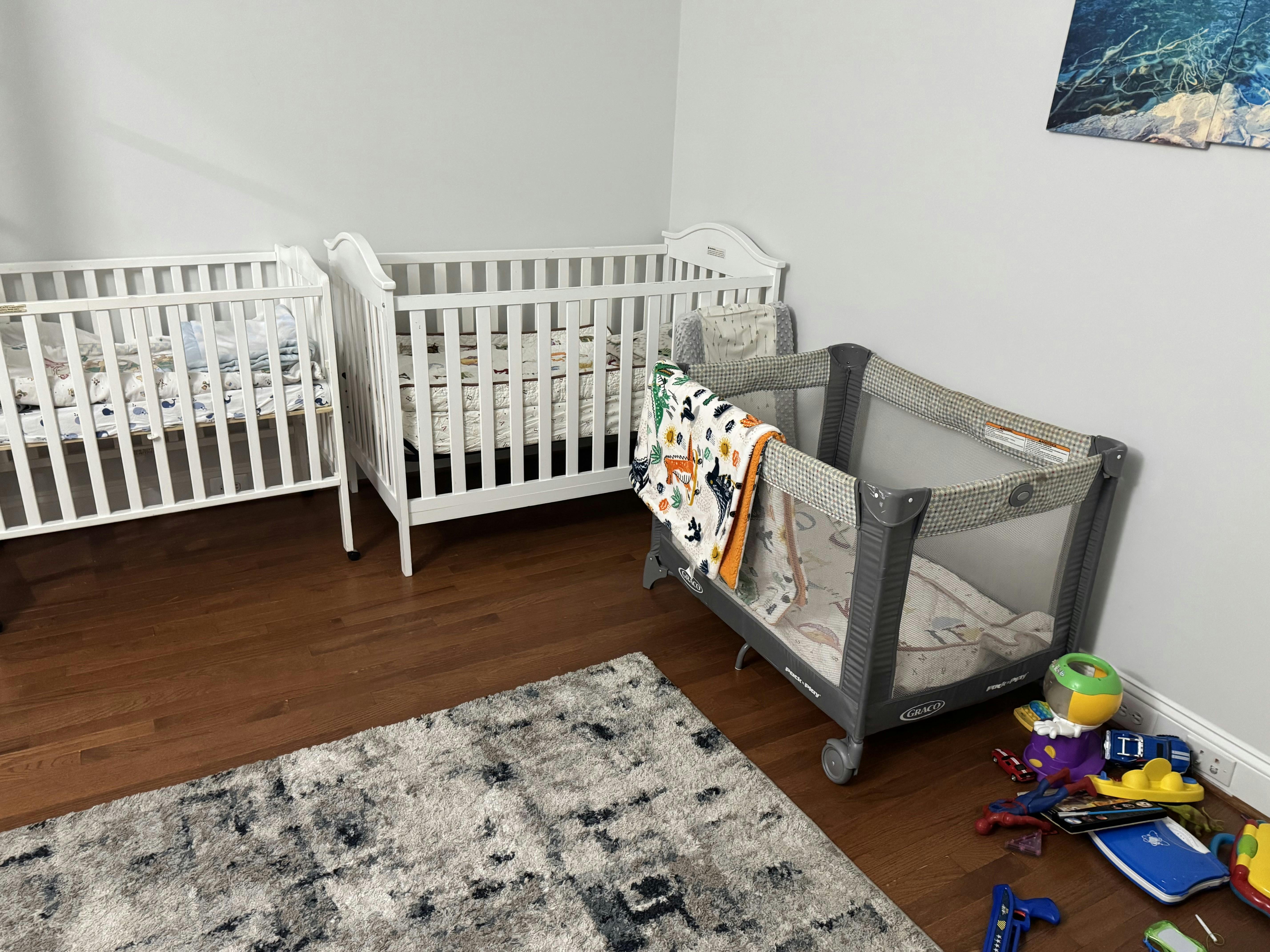 Two white cribs and a gray playard in a room